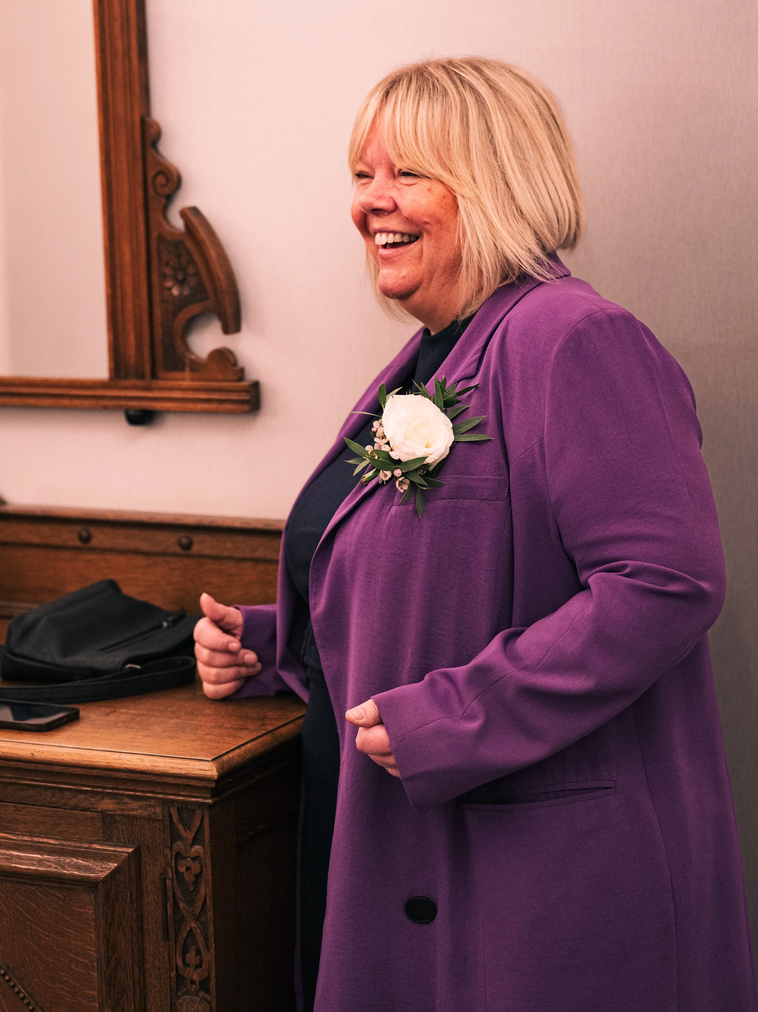 A woman with blonde hair smiling, wearing a purple blazer with a white flower boutonniere, standing near a wooden dresser.