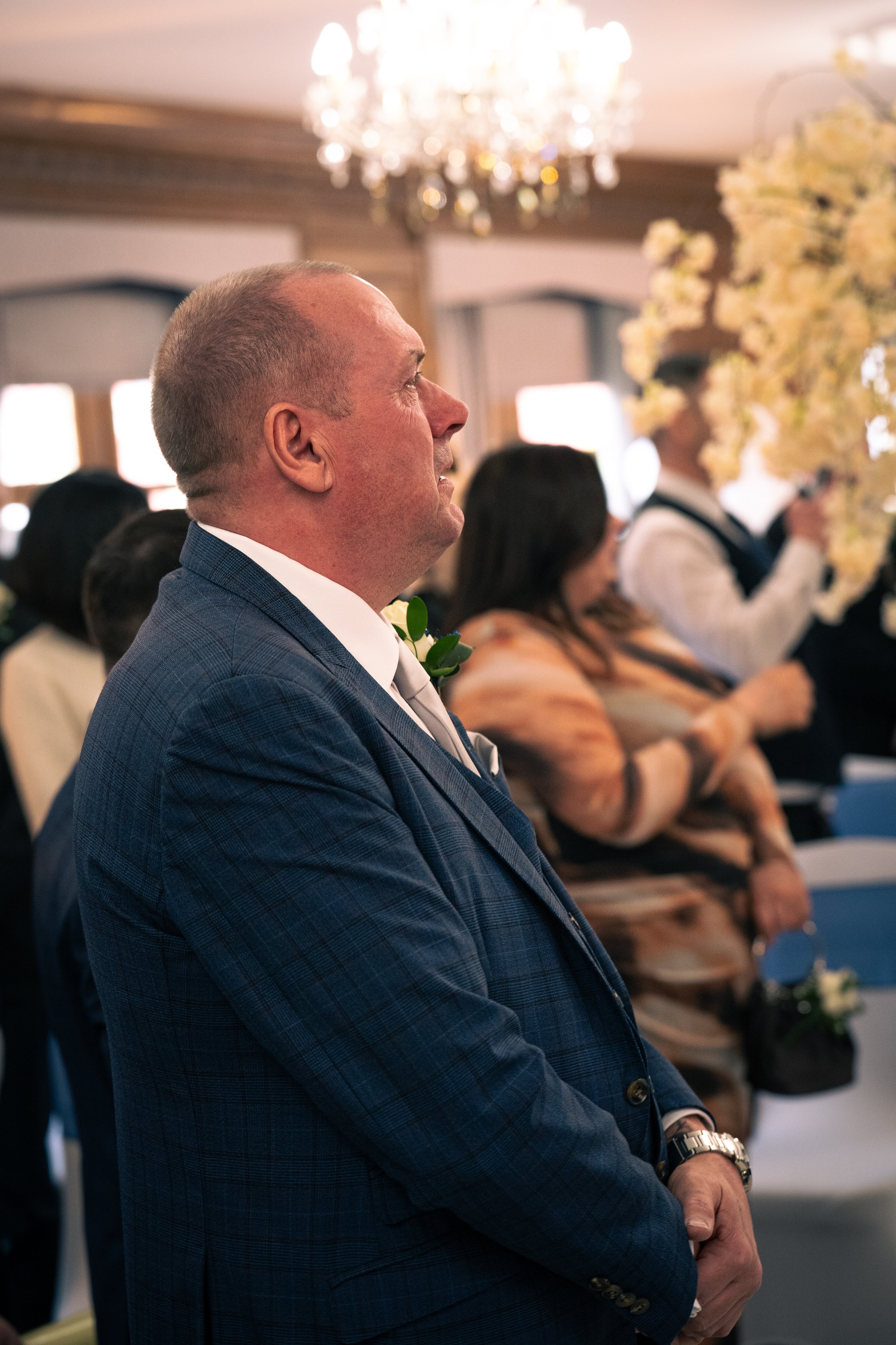 A man in a blue plaid suit and white shirt stands attentively at a formal event, with a boutonniere on his lapel, in a decorated room with chandeliers and floral arrangements.
