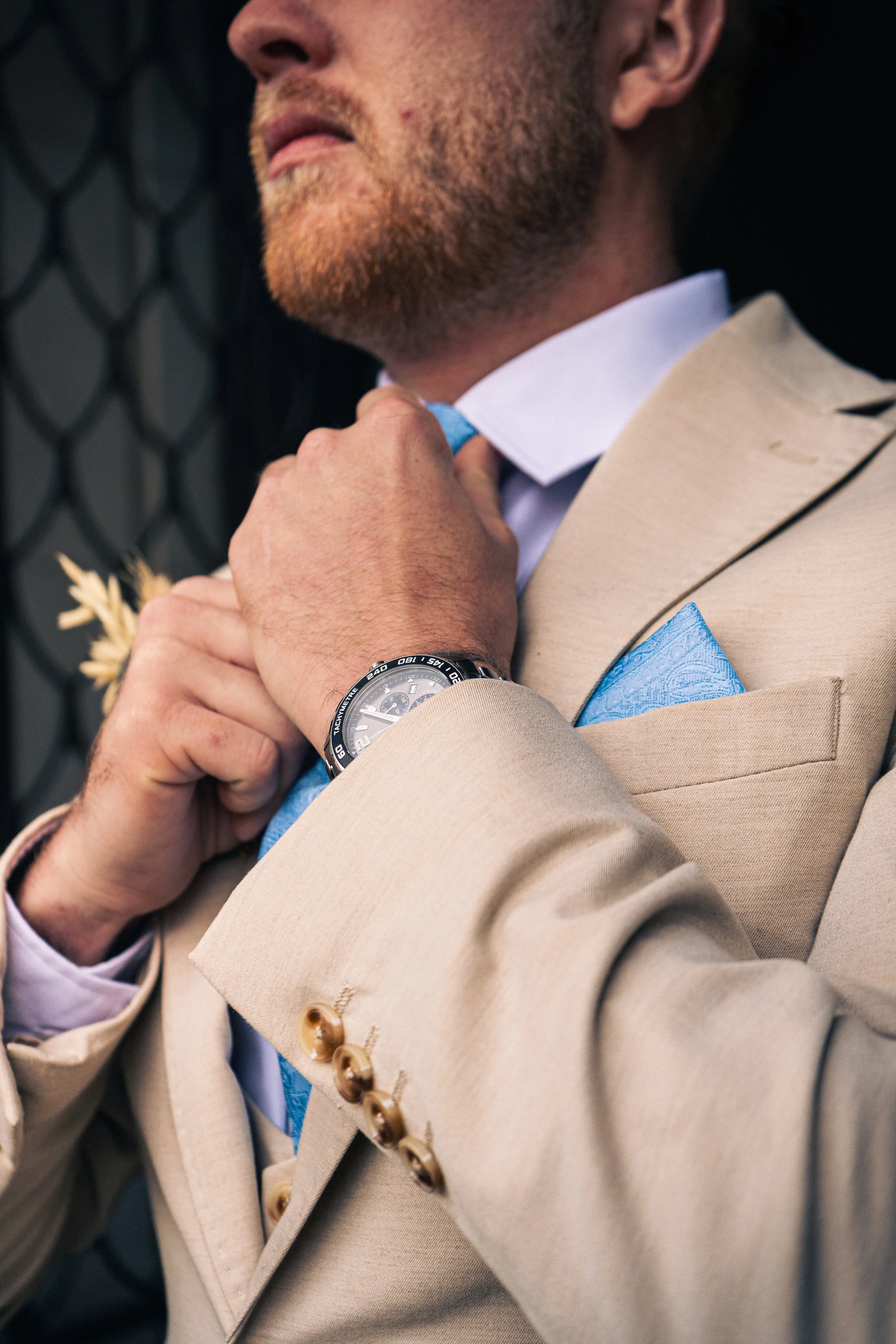 A man in a beige suit adjusting his blue tie, with a matching blue pocket square, wearing a wristwatch, and standing outdoors near a black metal fence.