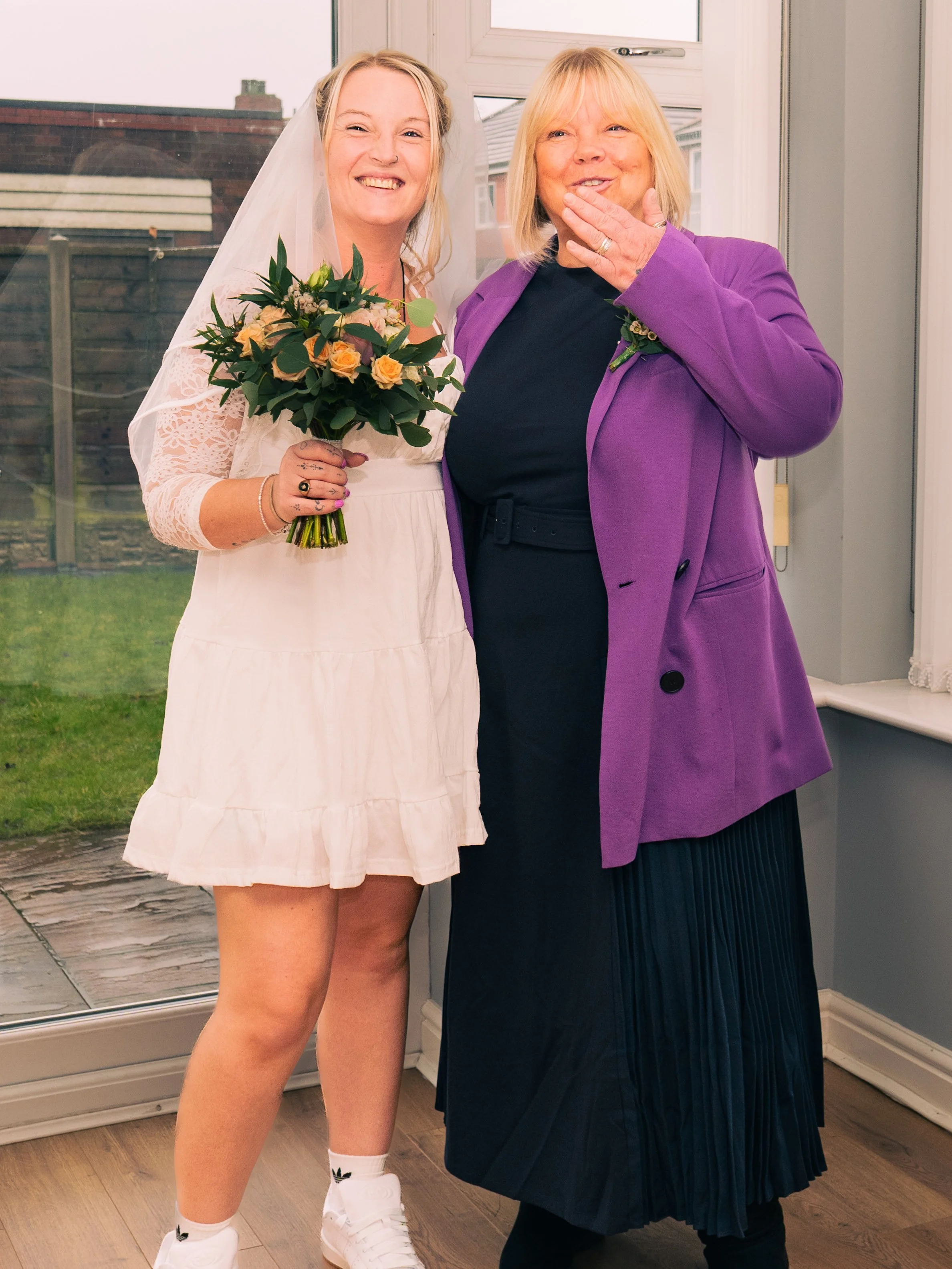 A smiling bride with a bouquet of flowers standing beside a woman covering her mouth and smiling, both inside a house with a glass door showing a backyard.