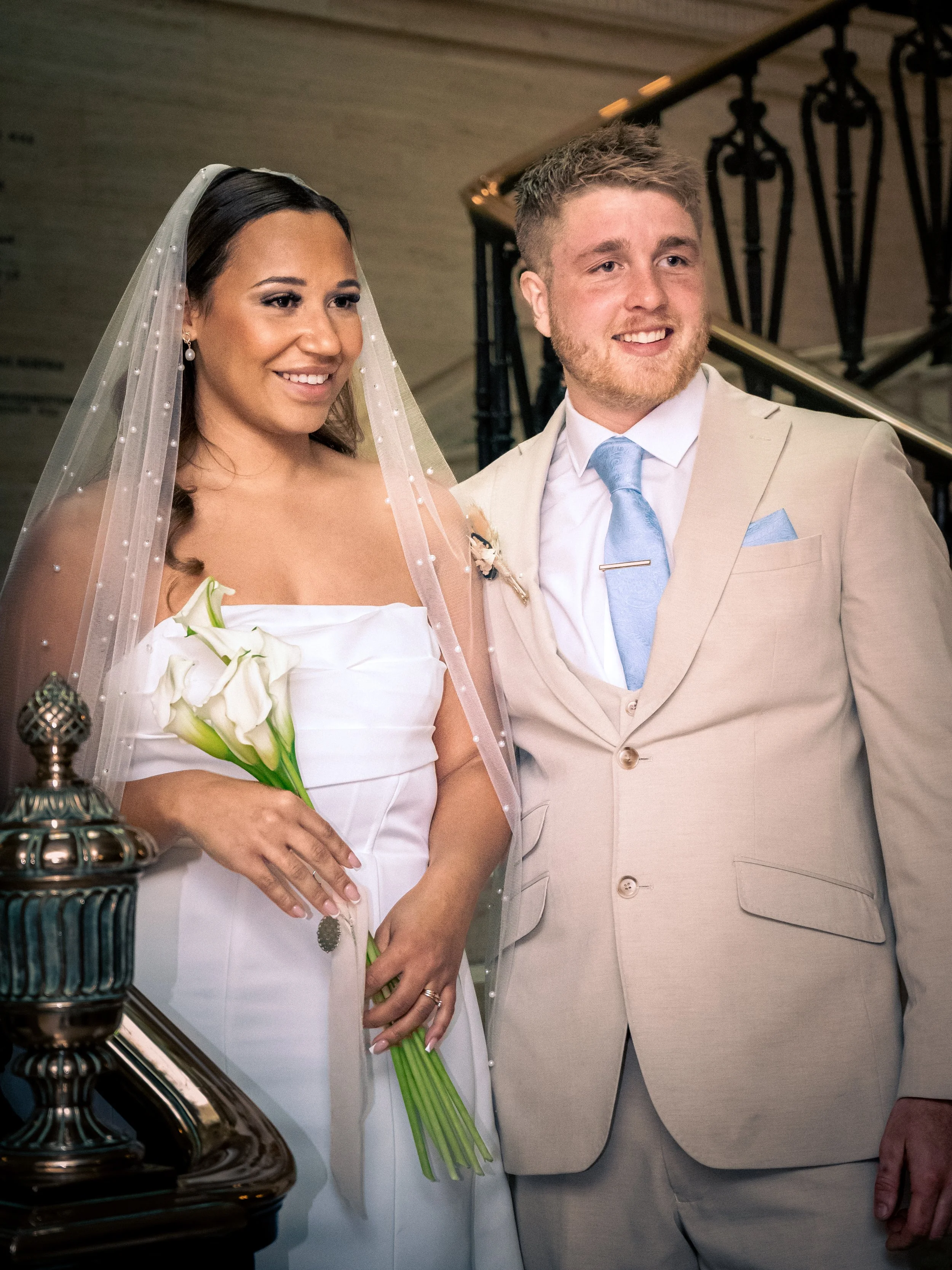A bride and groom standing together inside a building, smiling. The bride holds a bouquet of white flowers, and the groom is dressed in a light-colored suit with a blue tie.