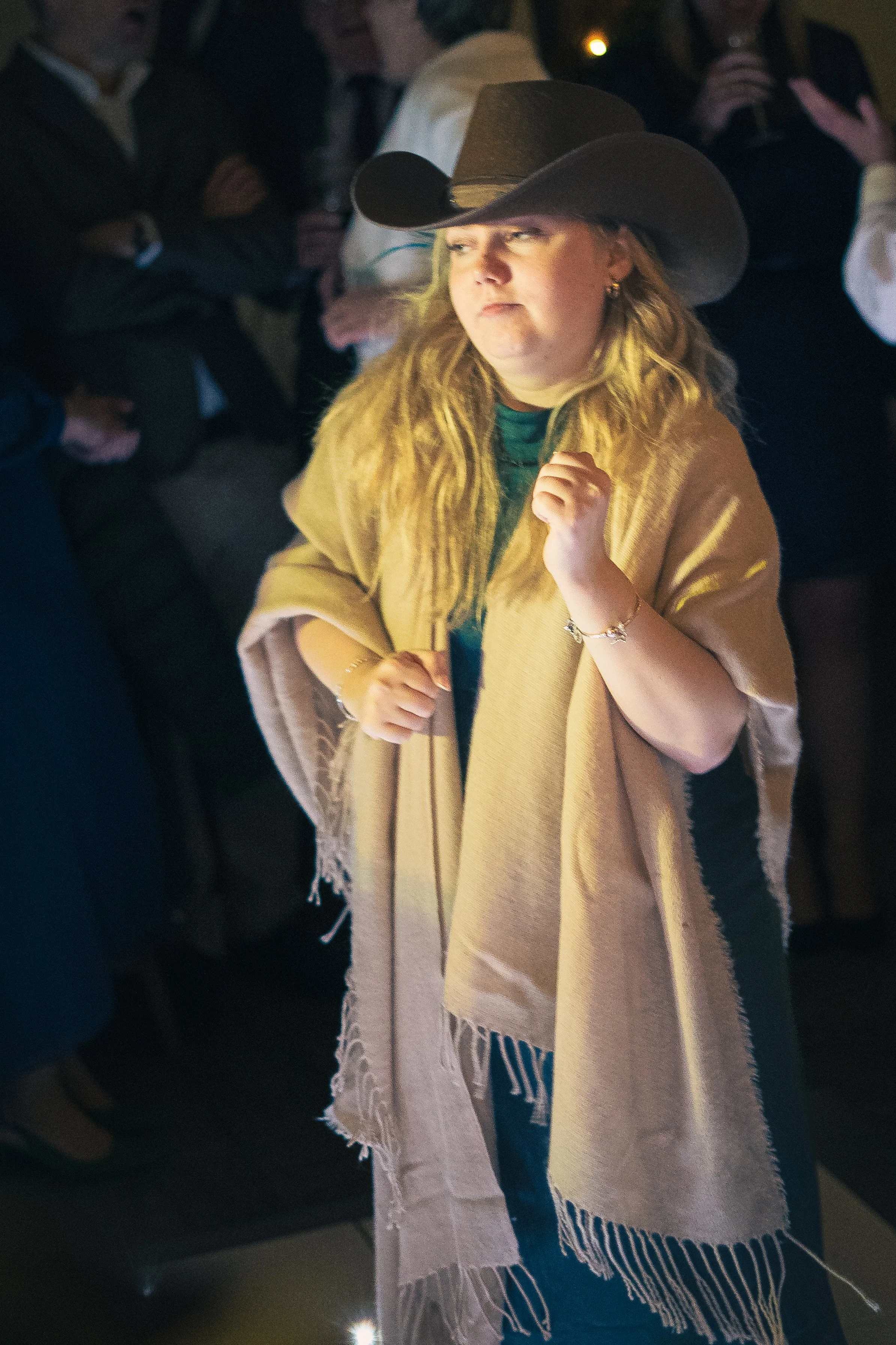 A woman wearing a large cowboy hat and a tan shawl stands in a crowd, looking to her left with a serious expression, in a dimly lit setting.