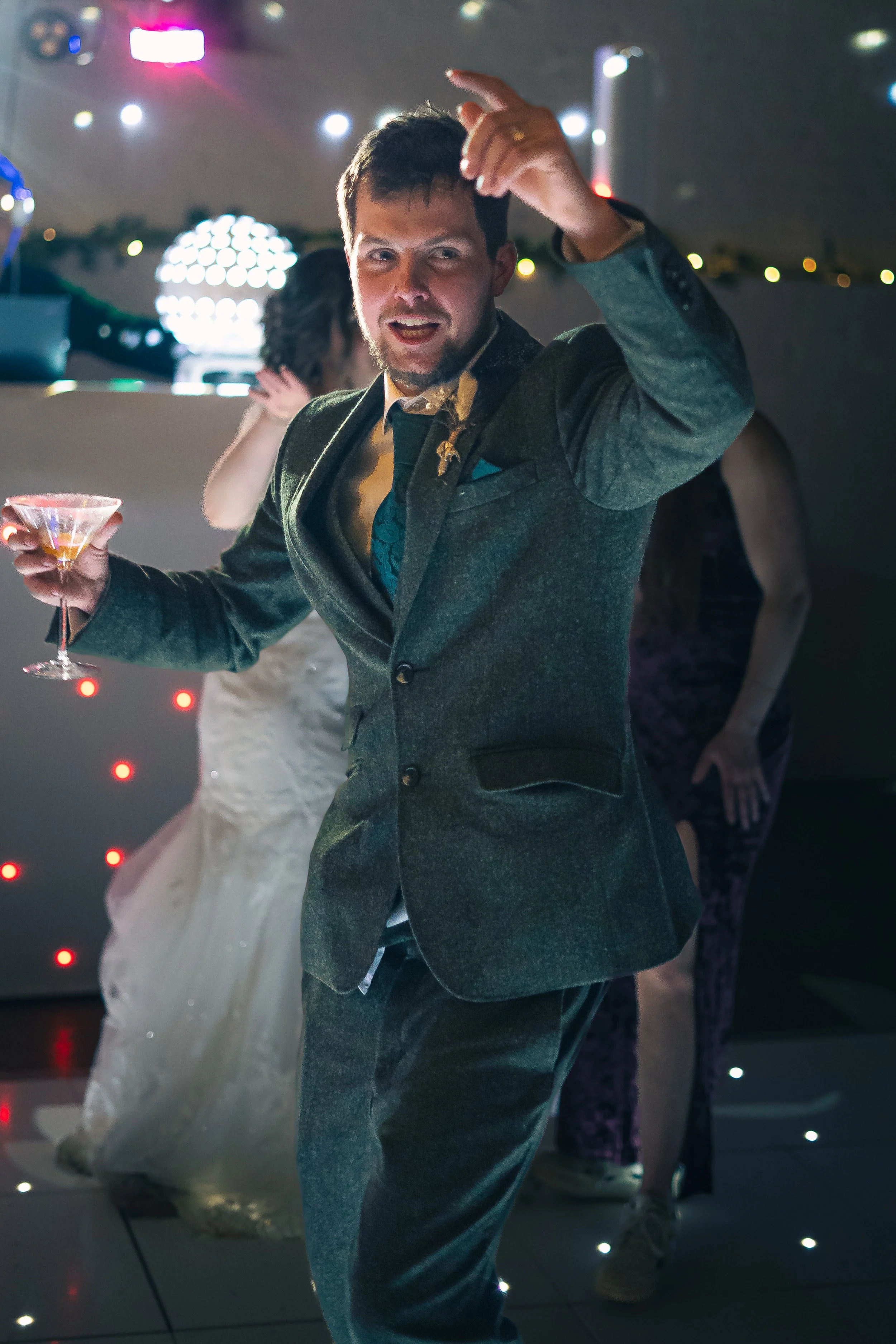 Man dancing at a celebration or party, holding a cocktail glass, with a woman in a wedding dress in the background and another woman nearby, all indoors with colorful lights.