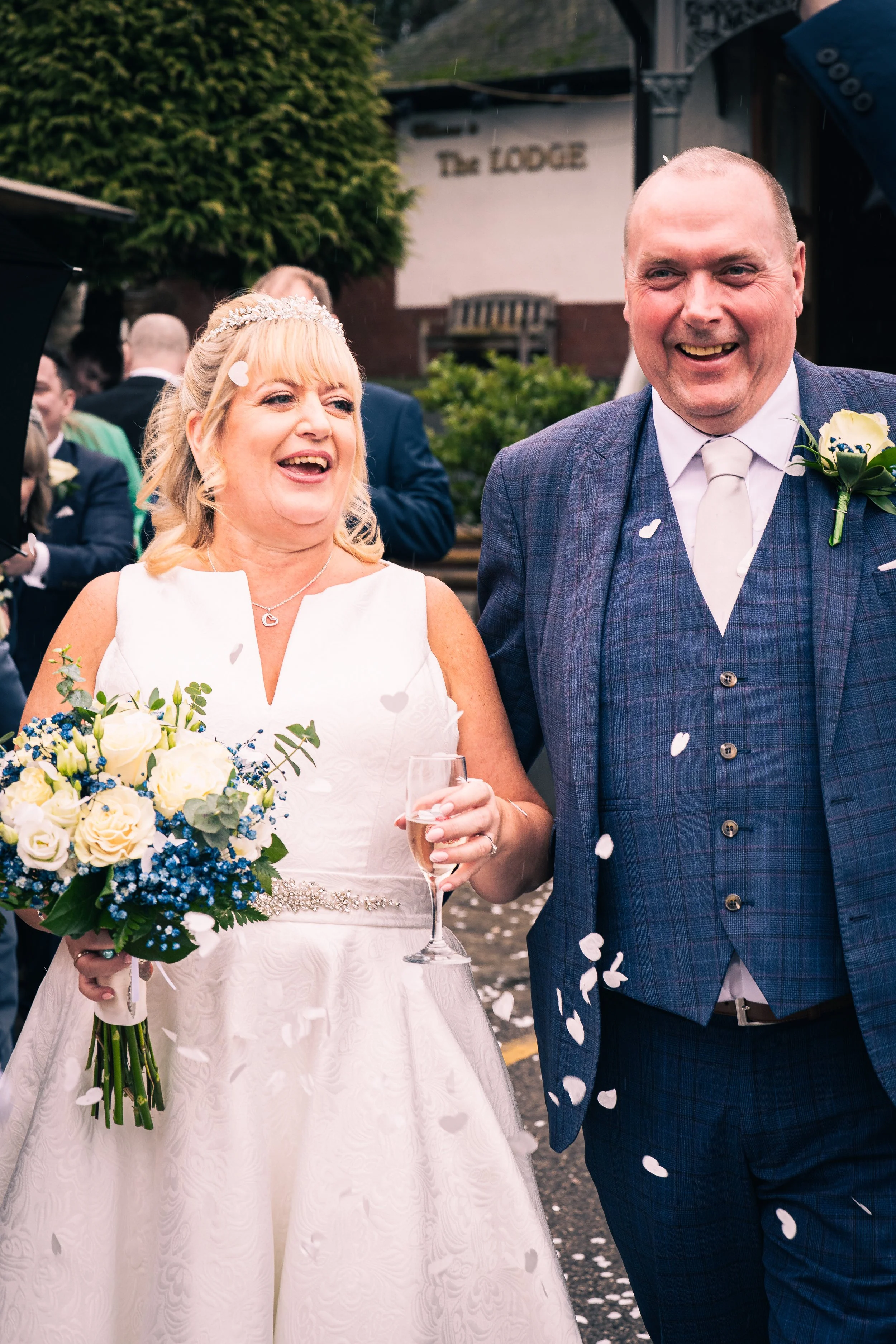 A bride and groom celebrating outside, surrounded by friends, with confetti falling around them. The bride is holding a bouquet of white and blue flowers and a glass of champagne, while the groom is dressed in a blue checkered suit and tie.
