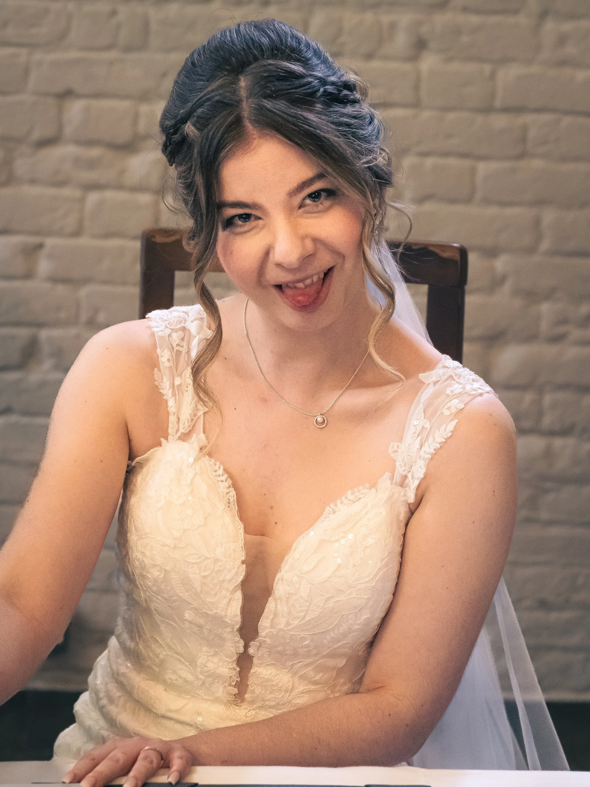 A young woman in a wedding dress sitting at a table, smiling with her tongue slightly out, in front of a brick wall.
