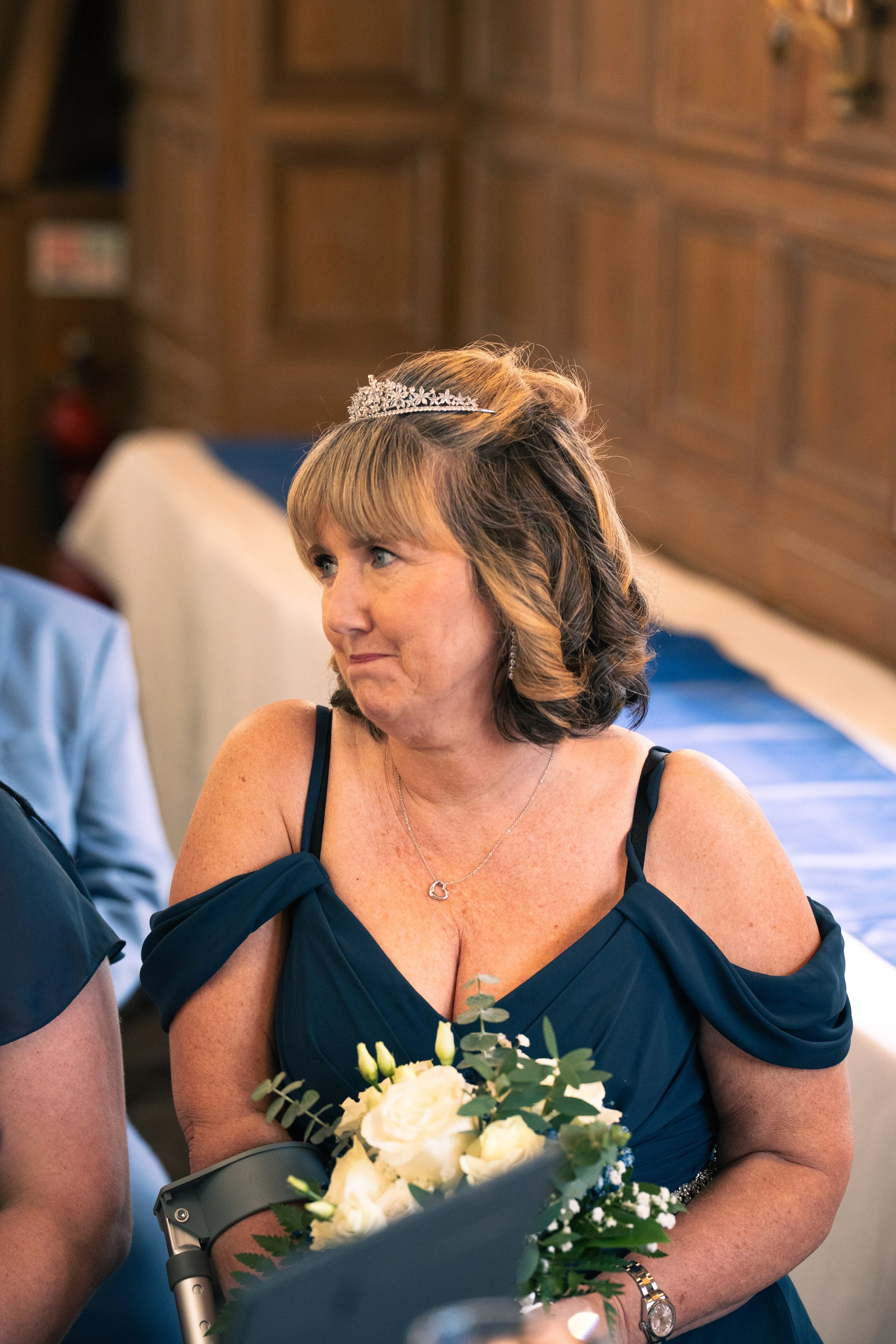 A woman wearing a blue off-shoulder dress and a tiara holding a bouquet of white flowers, attending a formal event.