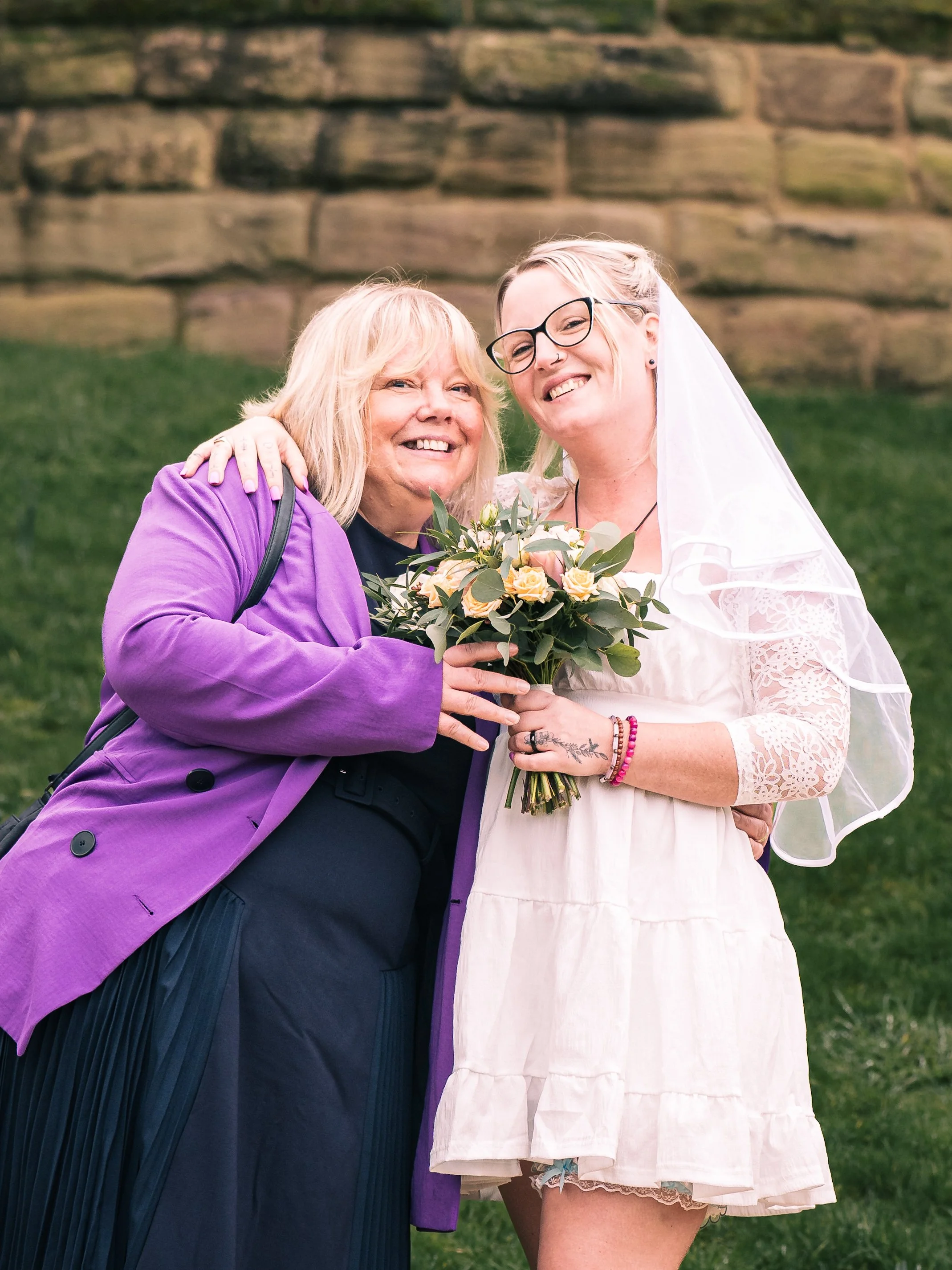 Two women smiling, one in a purple blazer and the other in a white dress with lace sleeves and a veil, holding a bouquet of flowers, standing outdoors in front of a stone wall.
