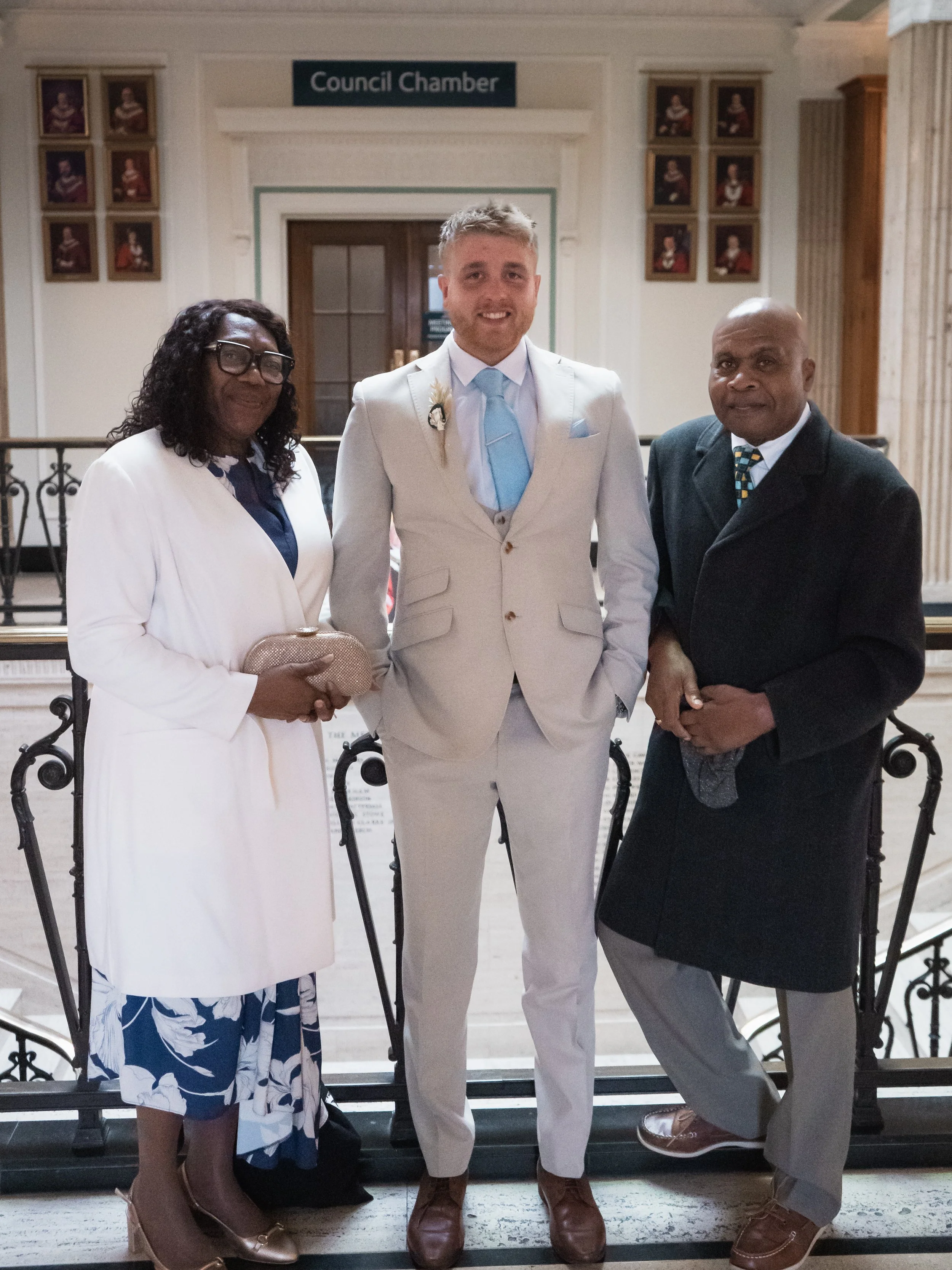 Three people dressed in formal attire standing in front of a sign that reads "Council Chamber" inside a building.