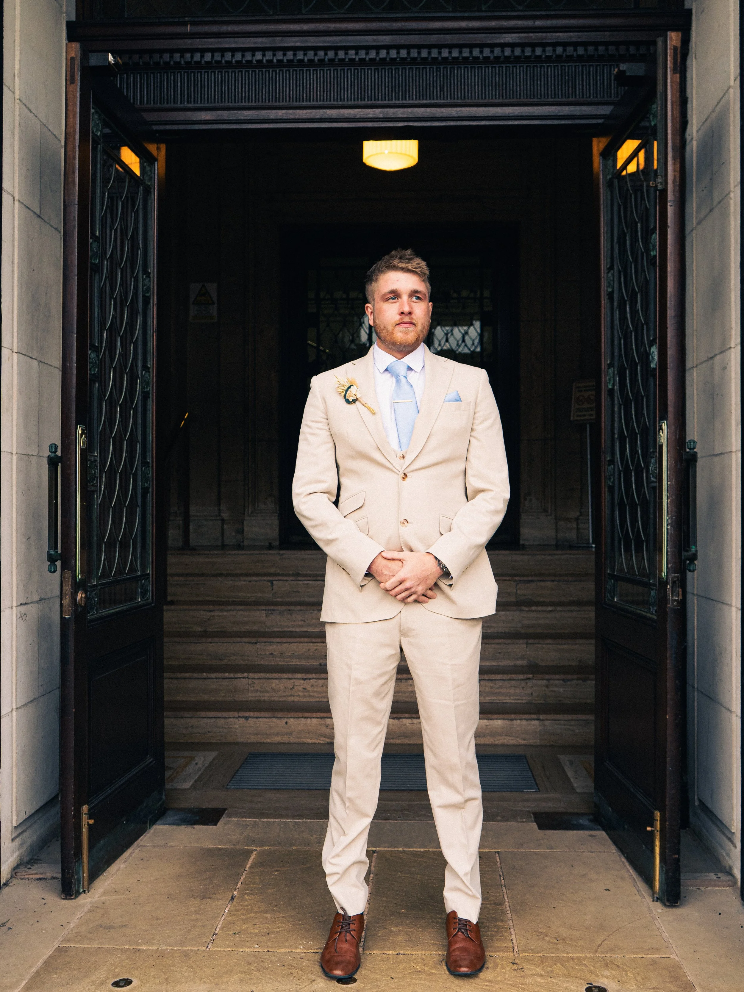 Man in cream suit with blue tie standing at open doorway, formal attire, wedding boutonniere, engaged in wedding or special occasion.