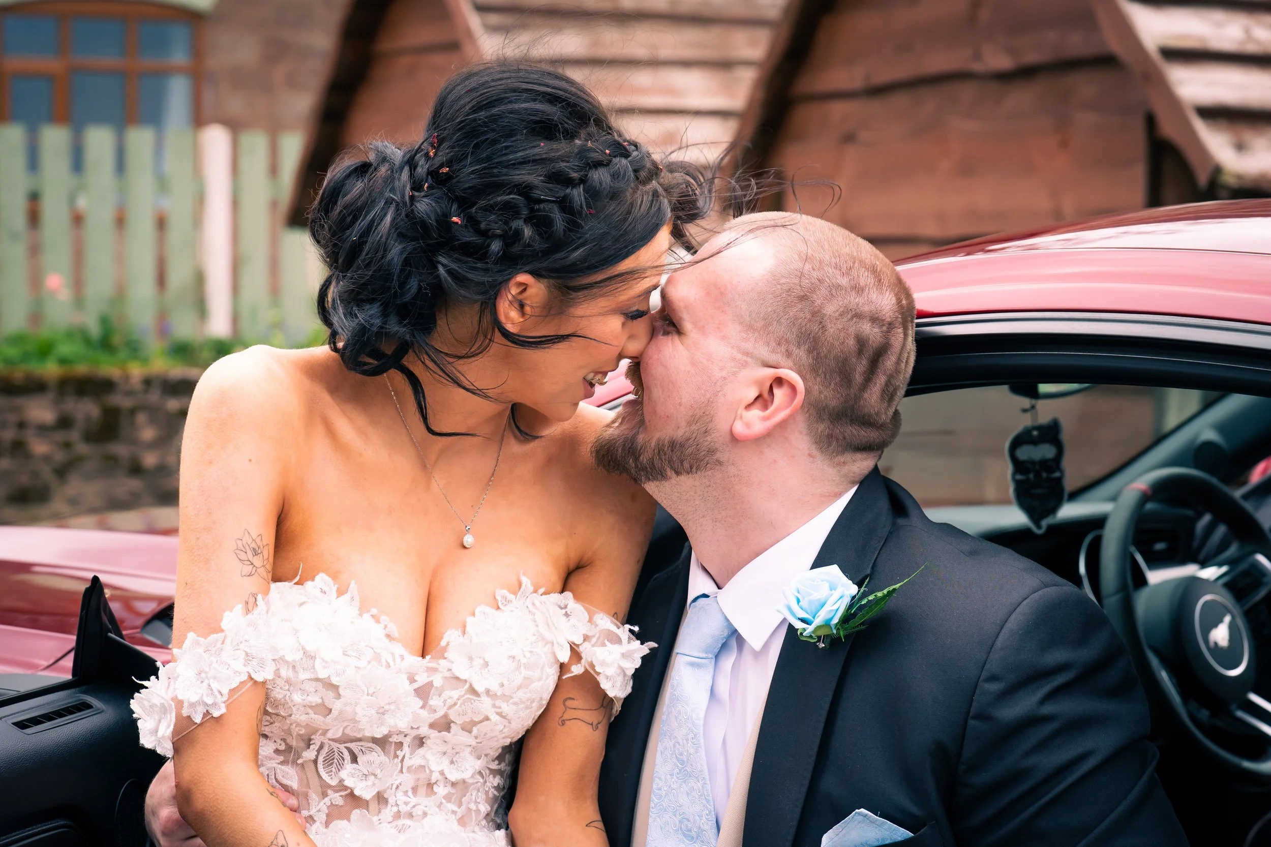 A newlywed couple sharing a kiss inside a red convertible car. The bride is wearing a white lace wedding dress with floral embroidery and has dark hair styled in braids with flower accents. The groom is wearing a black suit, white shirt, and light bl