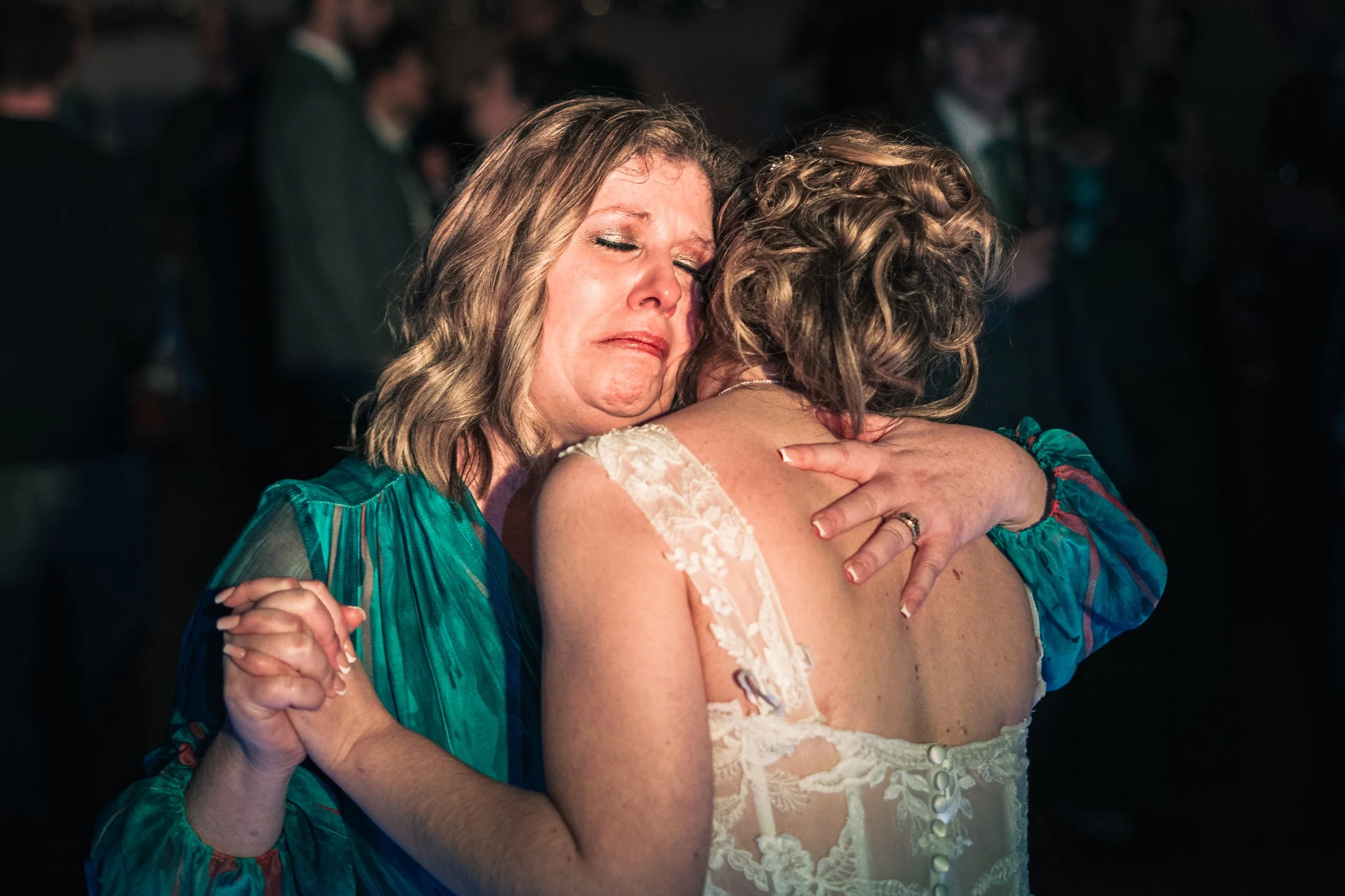Two women hugging at a wedding reception, one wearing a teal dress with sheer sleeves and the other in a wedding dress with lace straps, emotional moment.