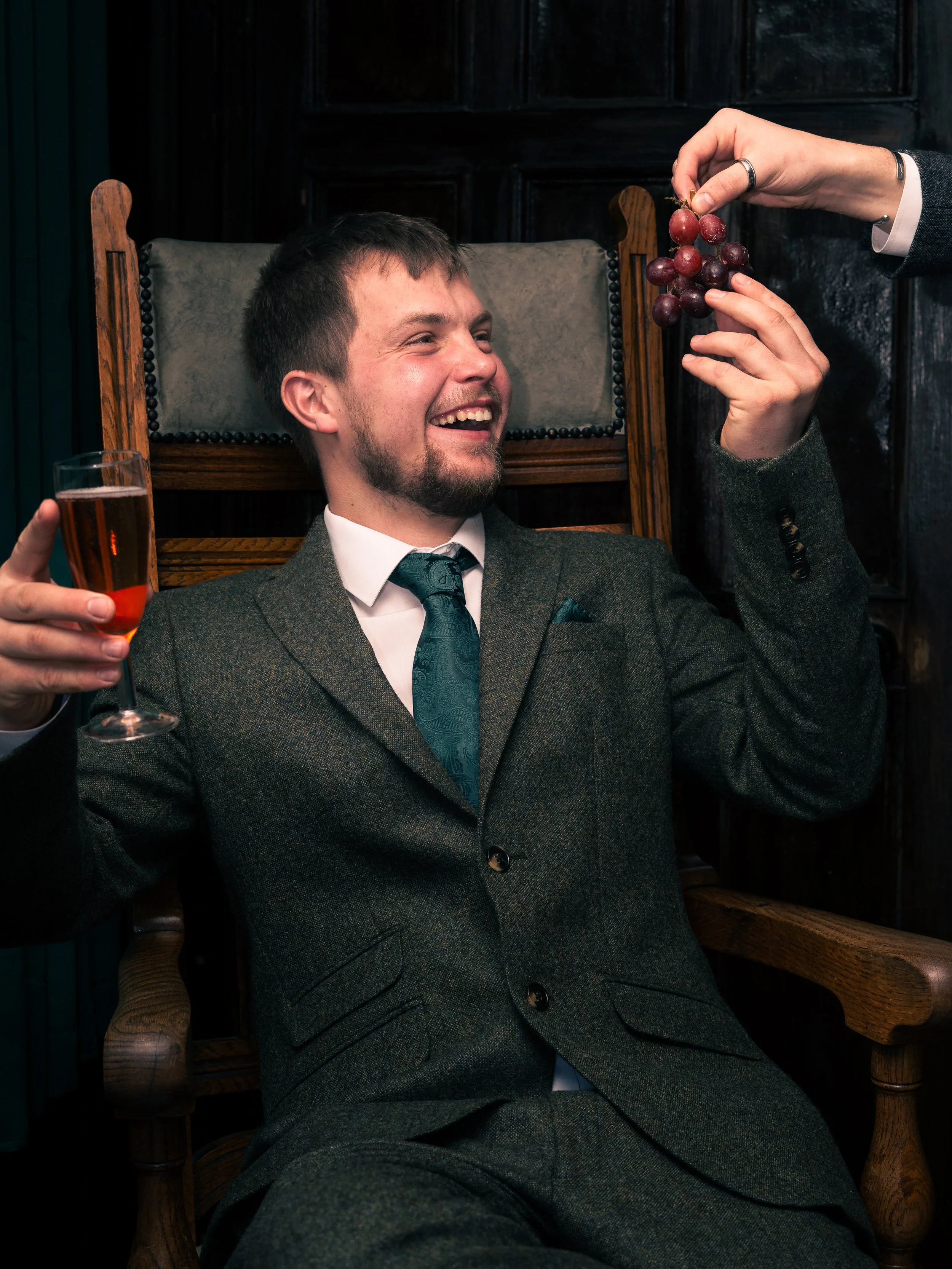 A man in a suit sitting on a wooden chair, holding a glass of rose-colored wine, smiling as another person holds a bunch of grapes above him.