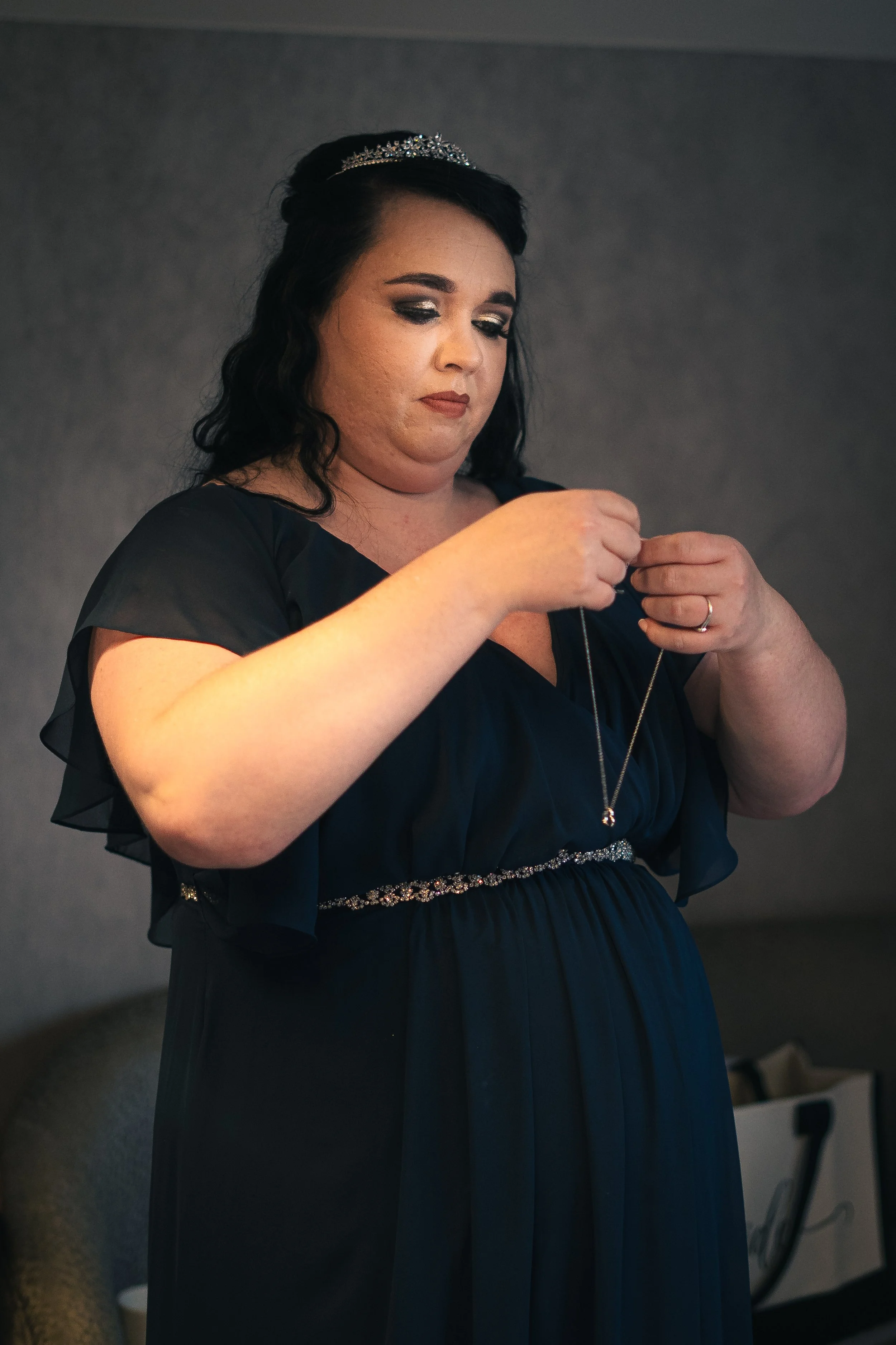 A woman in a navy blue dress with a tiara is putting on a necklace.