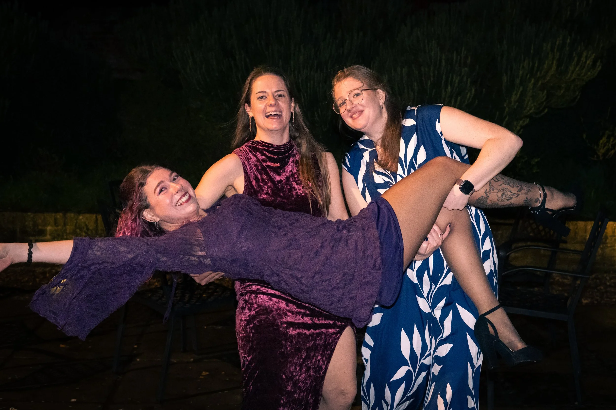 Three women smiling and having fun; one is being lifted horizontally by the other two at night outdoors.