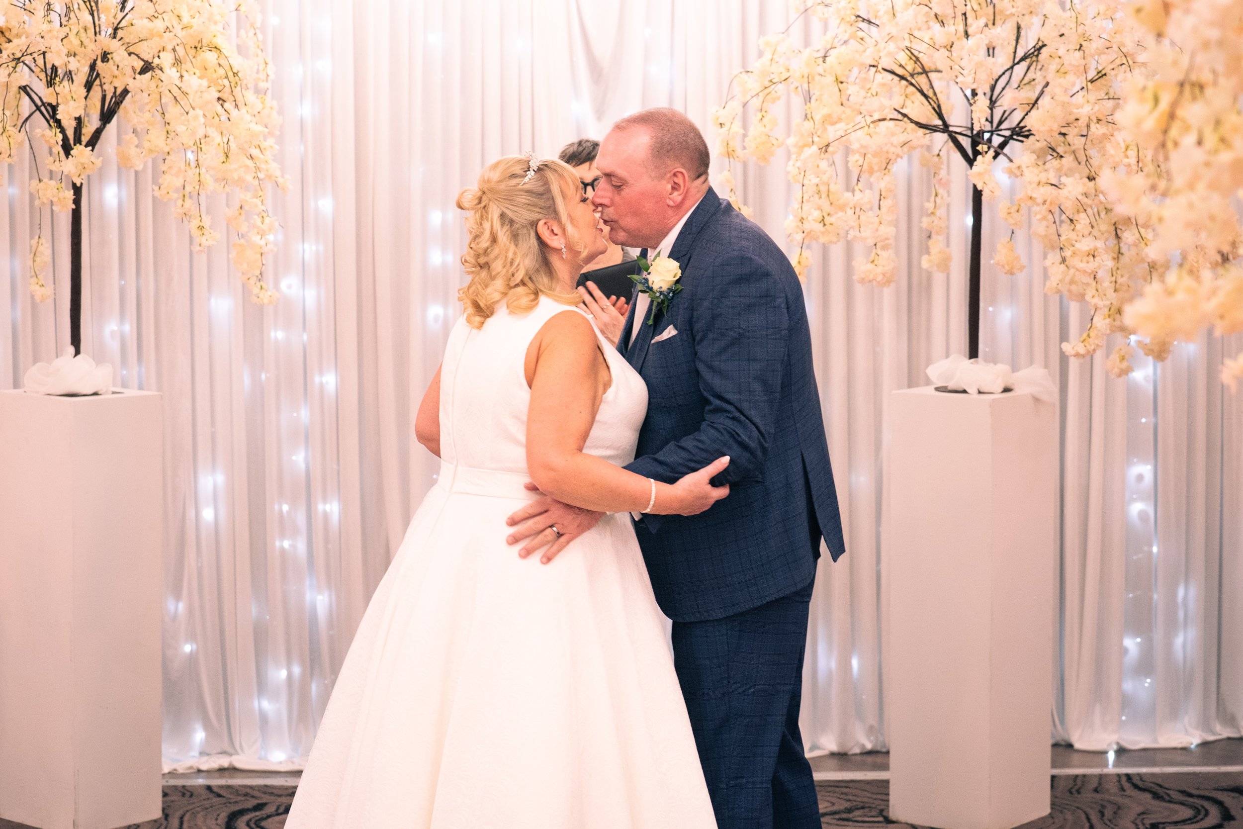 A bride and groom kiss during their wedding ceremony in front of a floral backdrop with white curtains and yellow flowers.