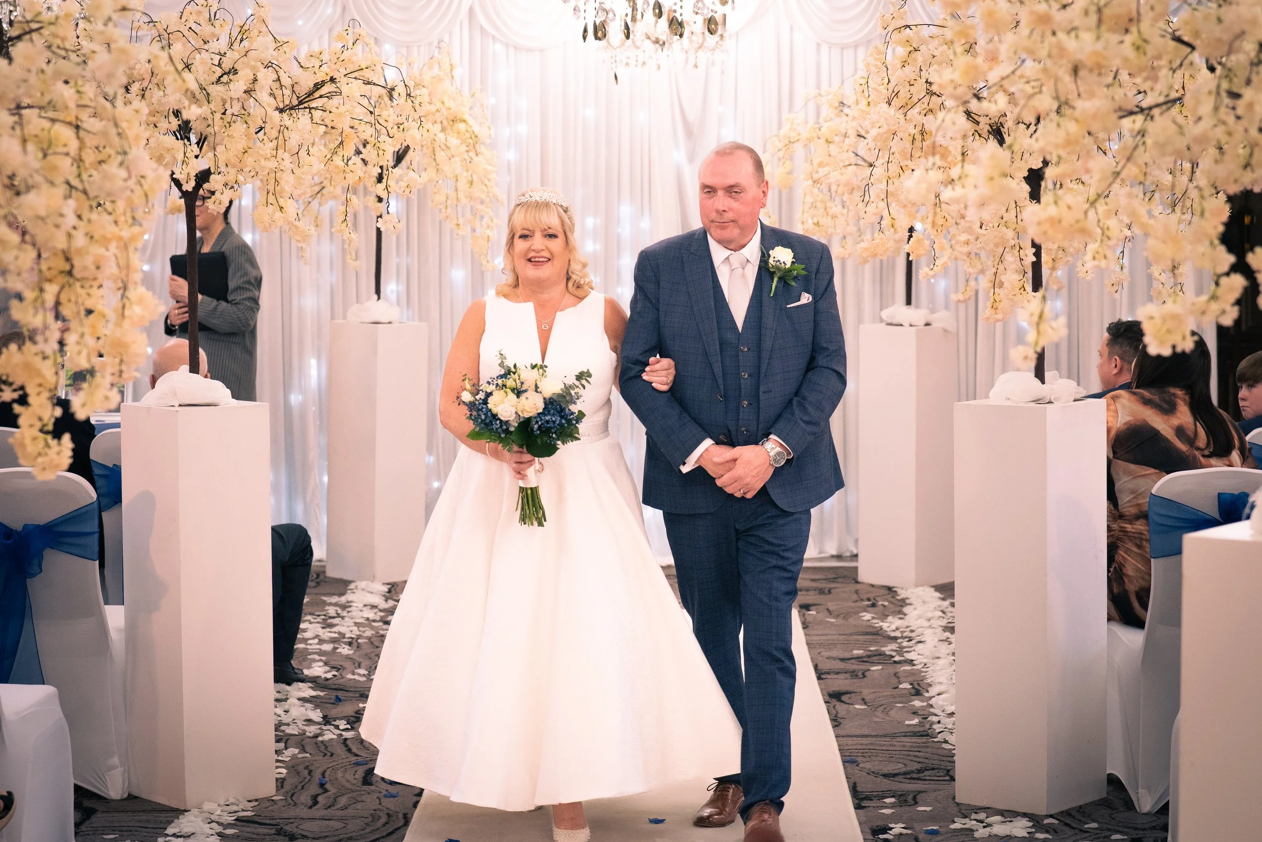 A bride in a white wedding dress holding a bouquet of white and blue flowers walking down the aisle with a man in a blue suit at a wedding ceremony. The ceremony is decorated with pink cherry blossom trees and white drapery.