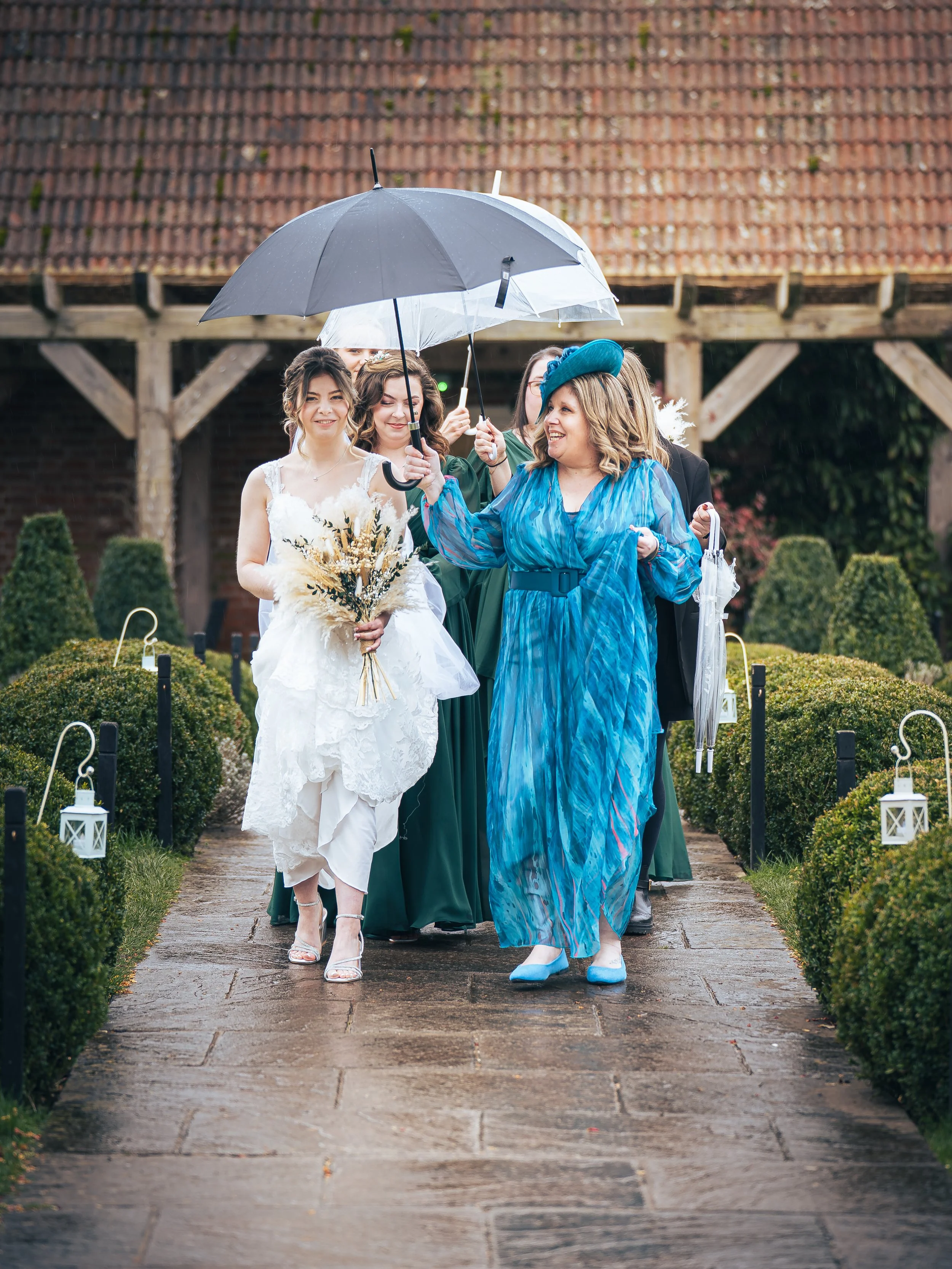 A group of women dressed in formal attire walking on a wet sidewalk, holding umbrellas, with one woman in a wedding dress, surrounded by greenery and lanterns along the path.