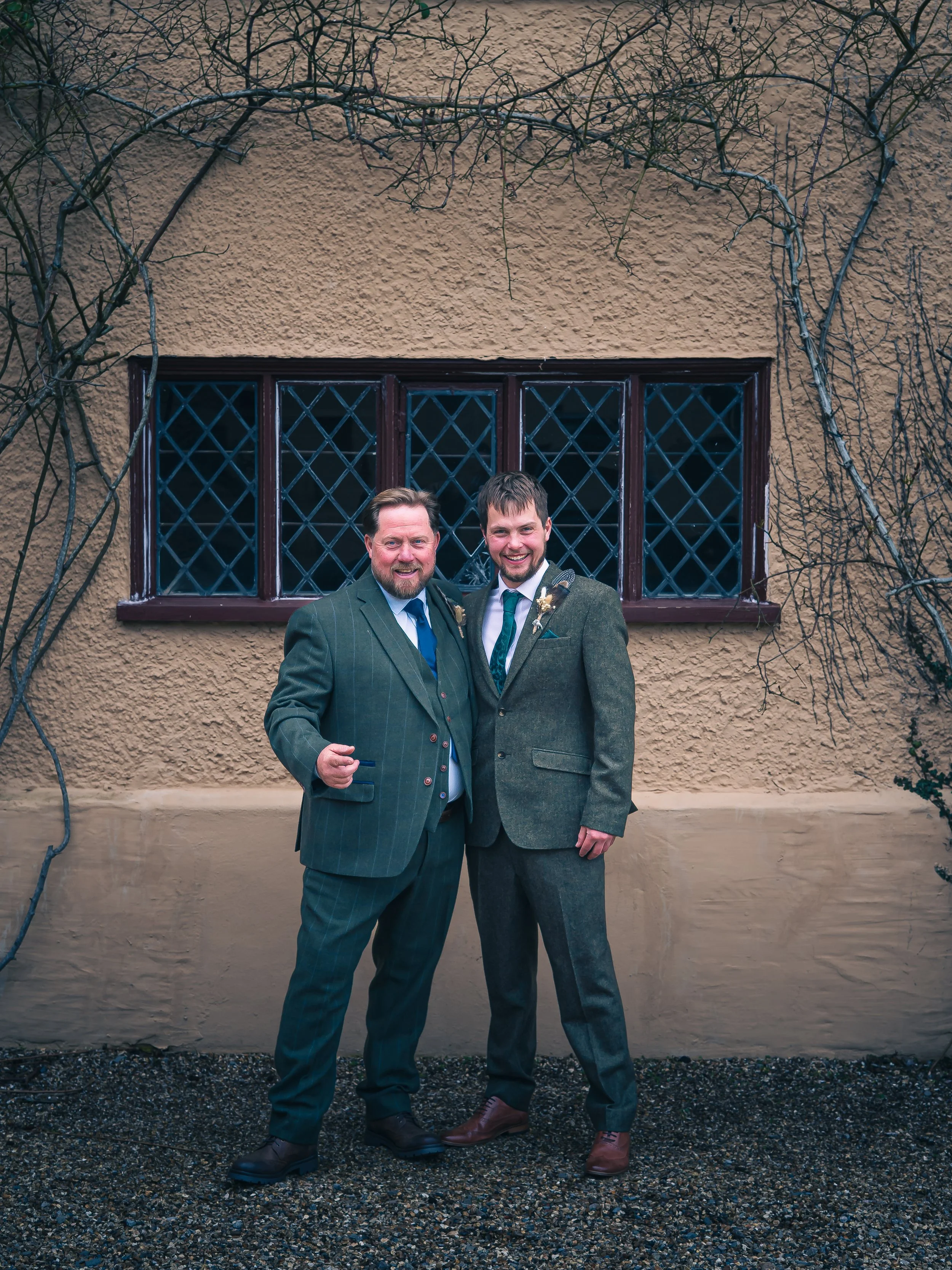 Two men in suits standing close together outdoors in front of a textured beige wall with a large window, smiling at the camera.