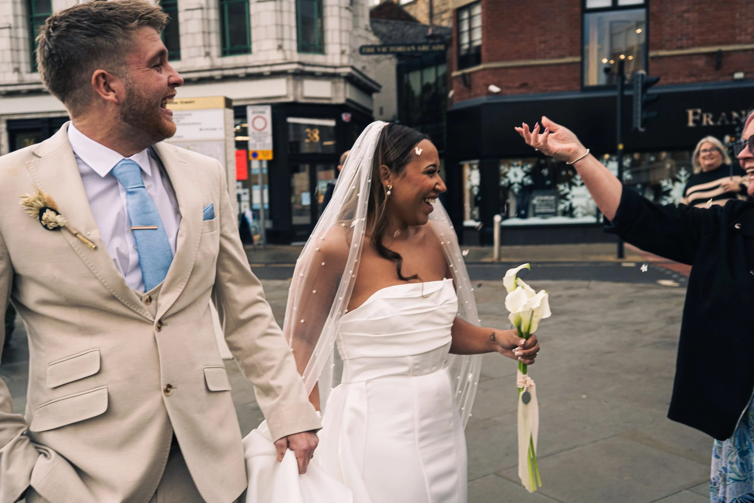 A wedding celebration with a bride in a white dress and veil holding a bouquet, standing next to a groom in a tan suit, smiling and holding hands, as a guest throws confetti in the air outdoors on a city street.
