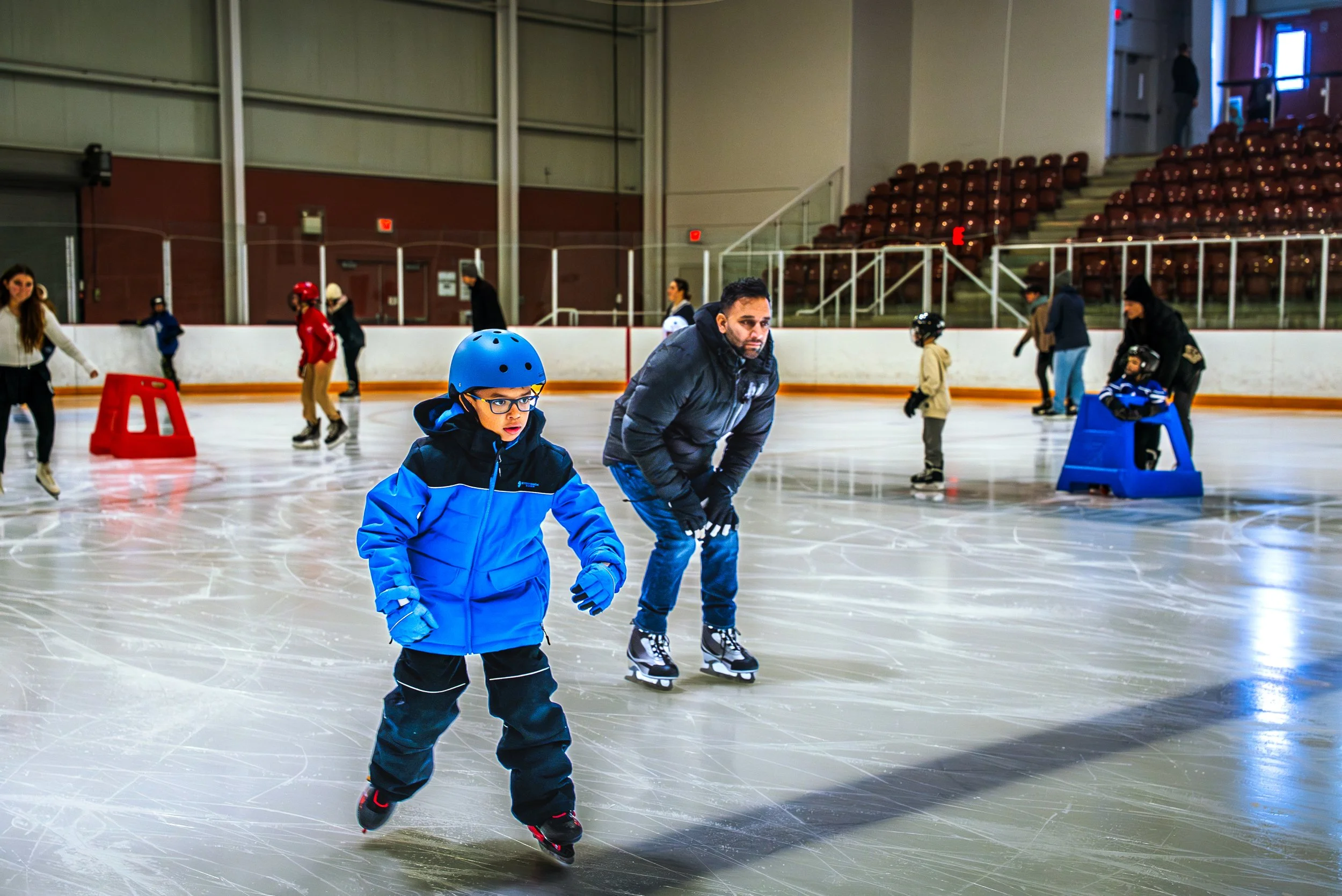 People ice skating at an indoor ice rink, including a young boy in a blue jacket and helmet, and an adult male skater nearby, with others skating and watching in the background.
