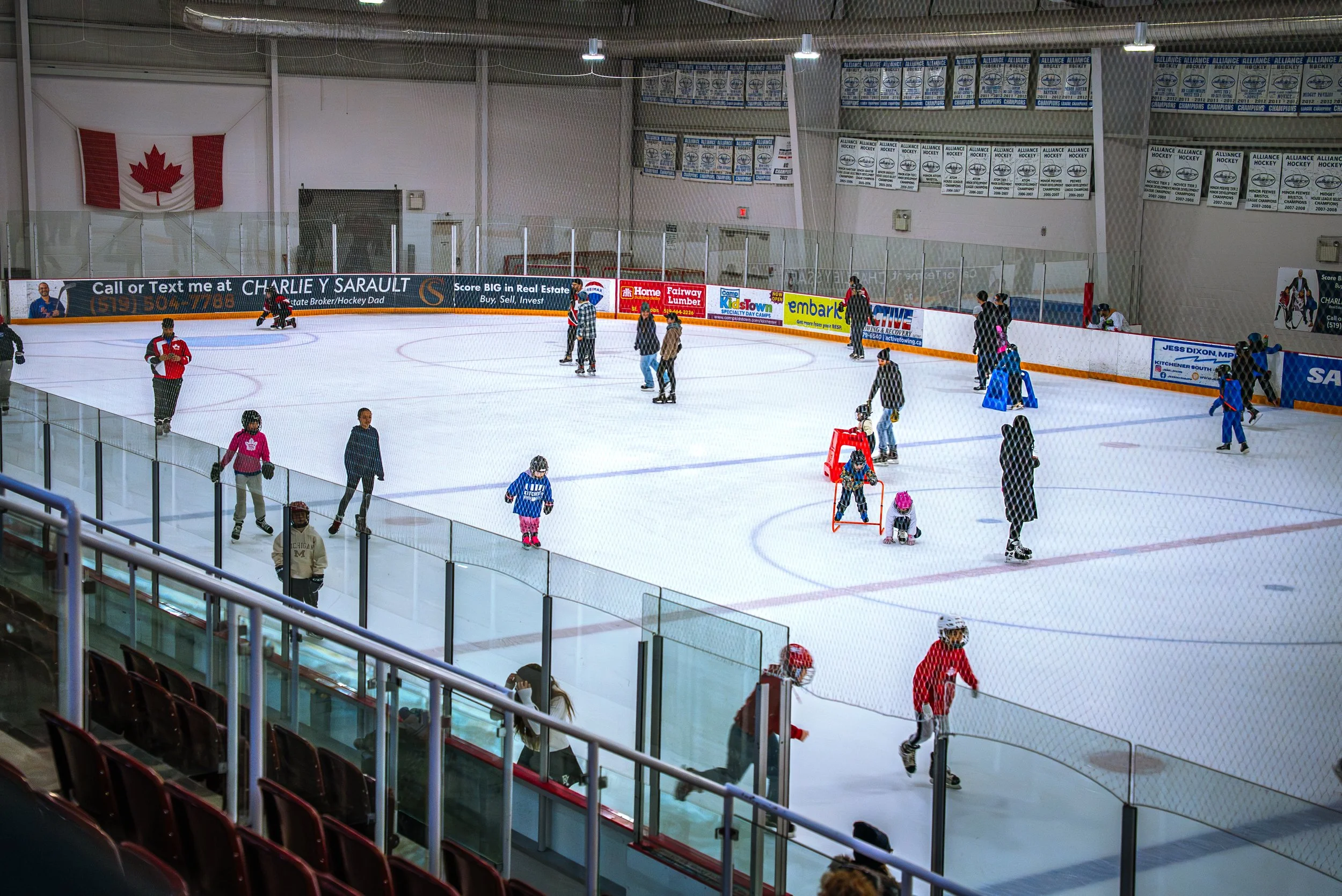People skating on an indoor ice hockey rink, with some children using training aids, and a large Canadian flag hanging on the wall.