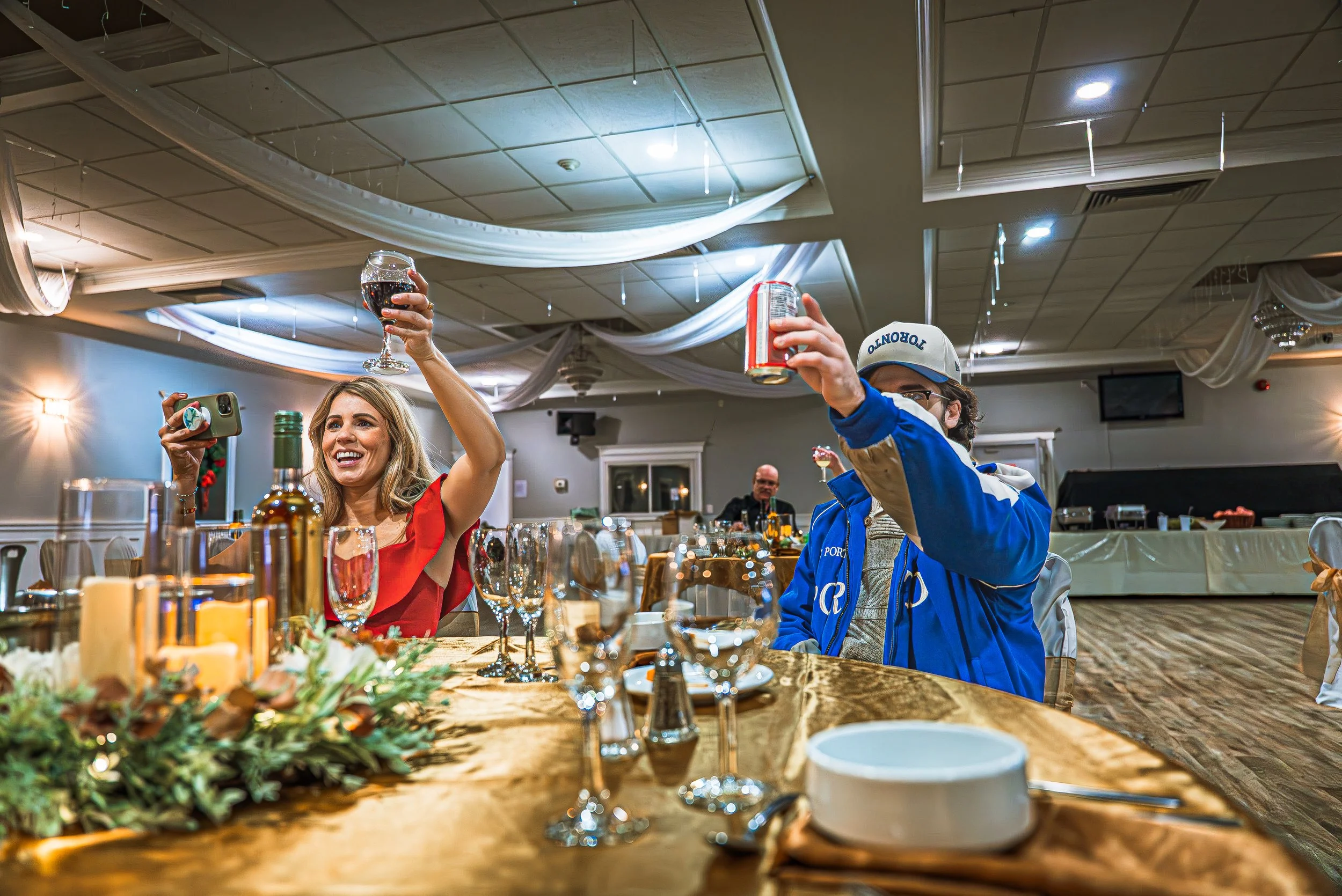 People celebrating at a banquet table indoors, raising glasses for a toast, with decorated ceiling and table setting including wine glasses, bottles, and a festive centerpiece.