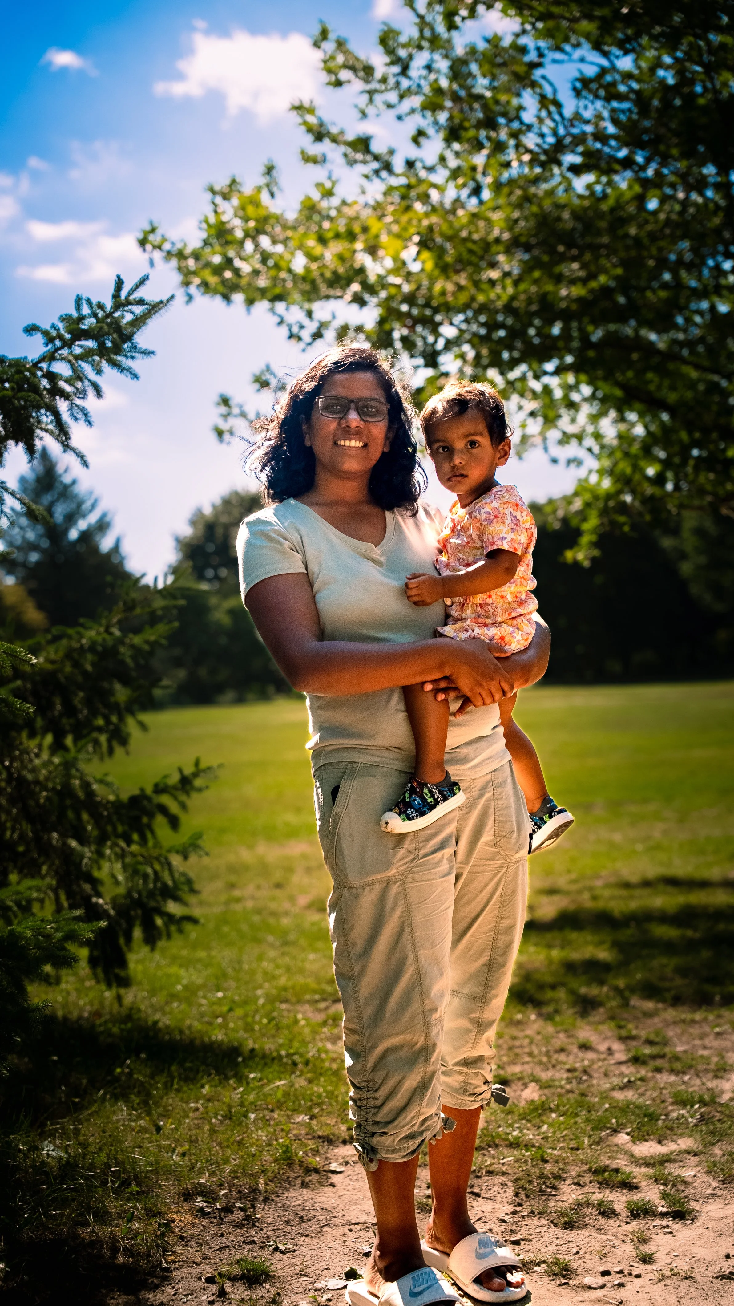 A woman with dark curly hair and glasses holding a young girl with dark hair dressed in a patterned shirt and shorts outdoors on a sunny day, with trees and a blue sky with clouds in the background.