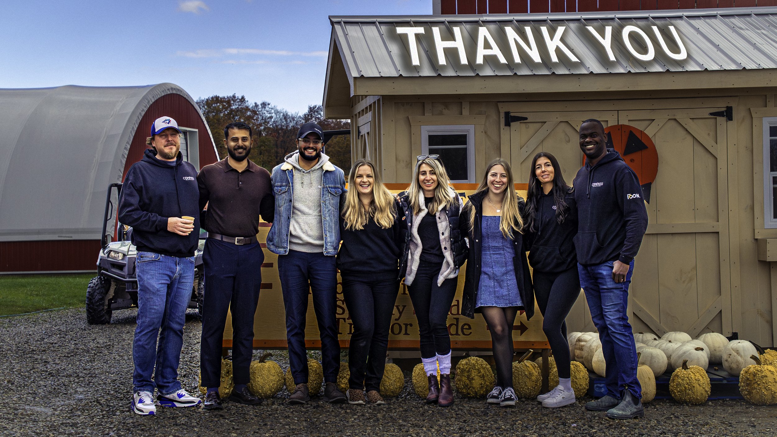 Group of seven diverse people smiling and standing together outdoors in front of a small wooden building with a "Thank You" sign, pumpkins, and hay bales on display.