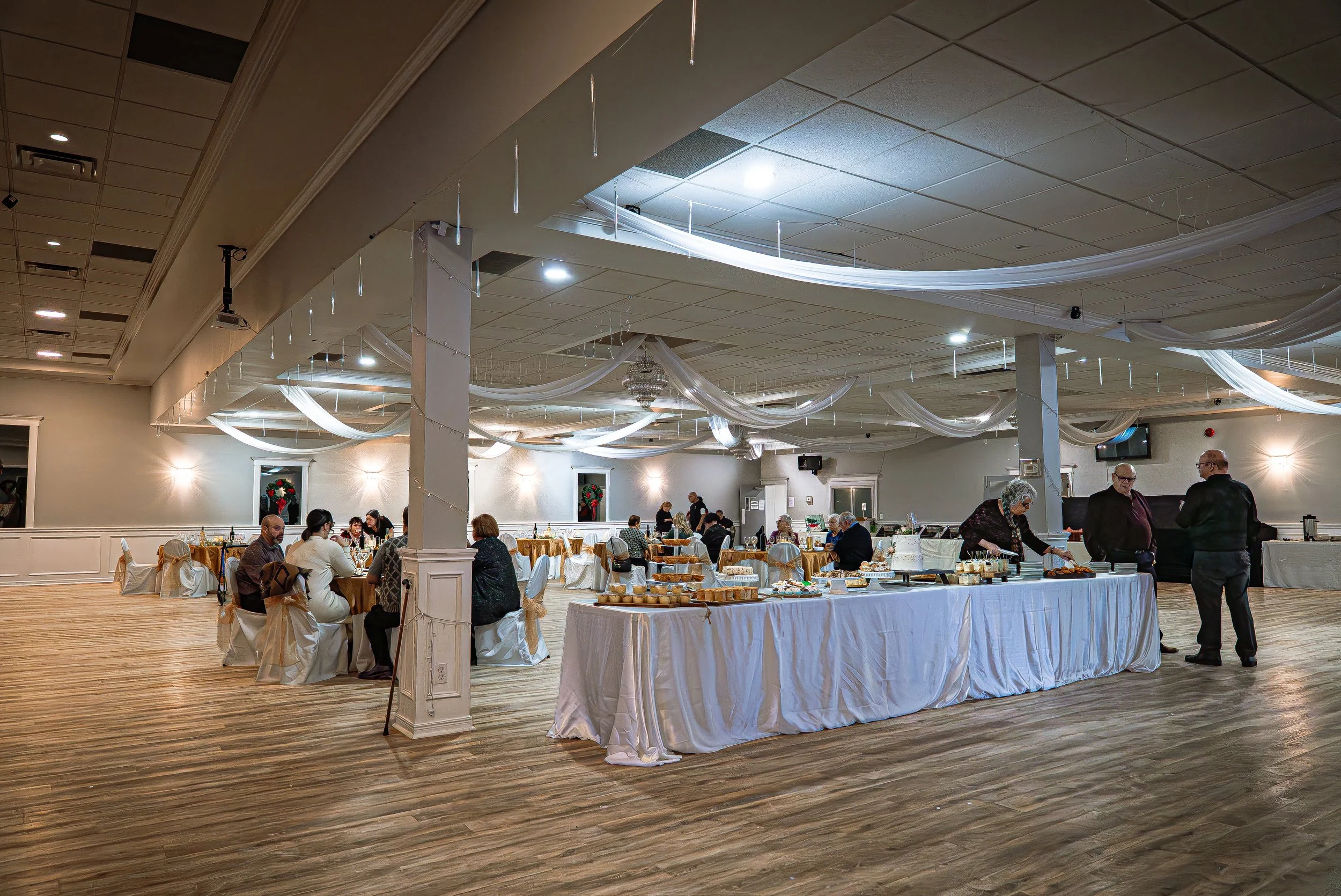 A banquet hall with decorated ceiling, round tables with guests, and an extended table of desserts in the foreground.