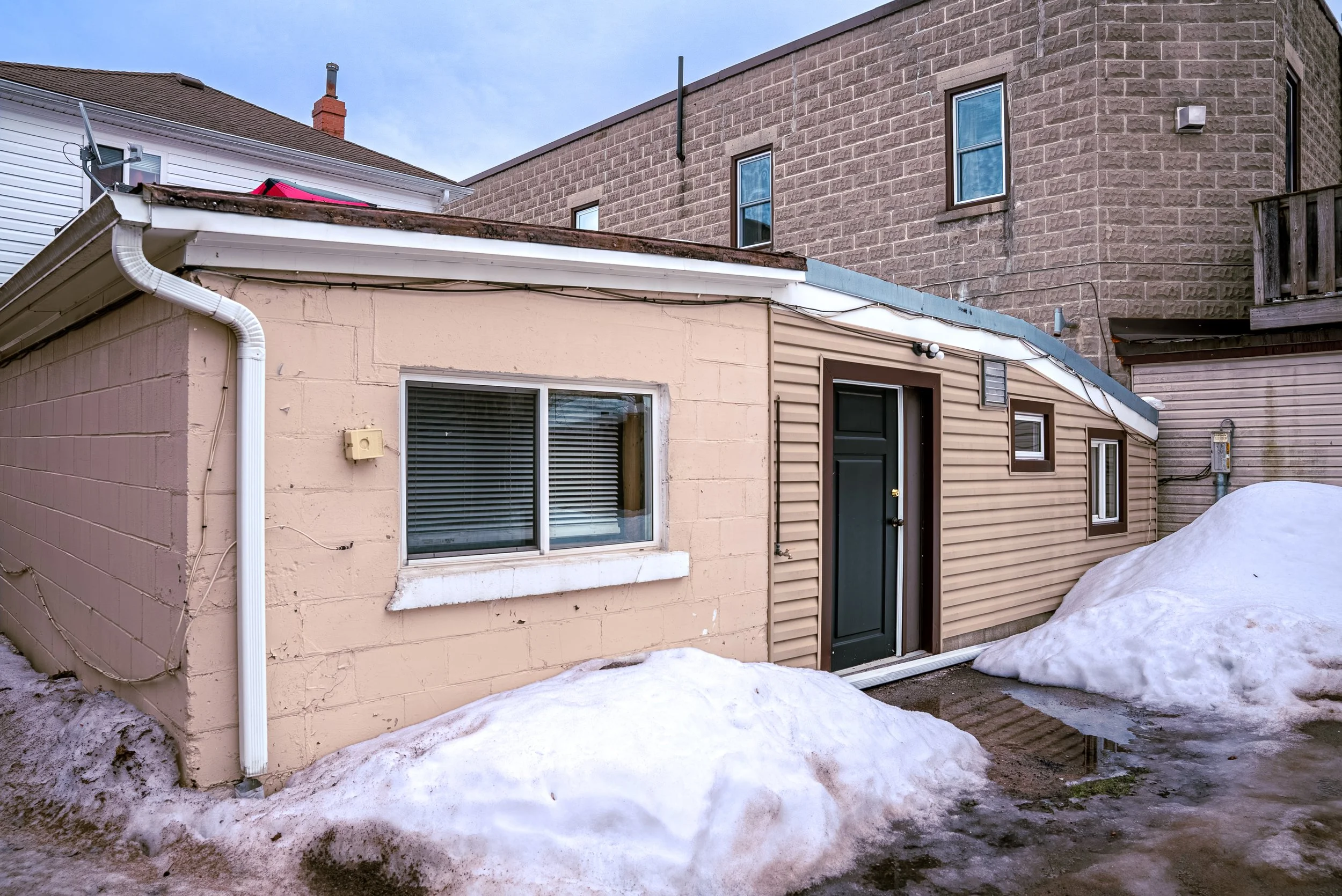Backyard view of a house with snow on the ground, showing beige and brown exterior walls, a sliding glass door, and windows. The backyard appears icy and wet, with some snow accumulation.