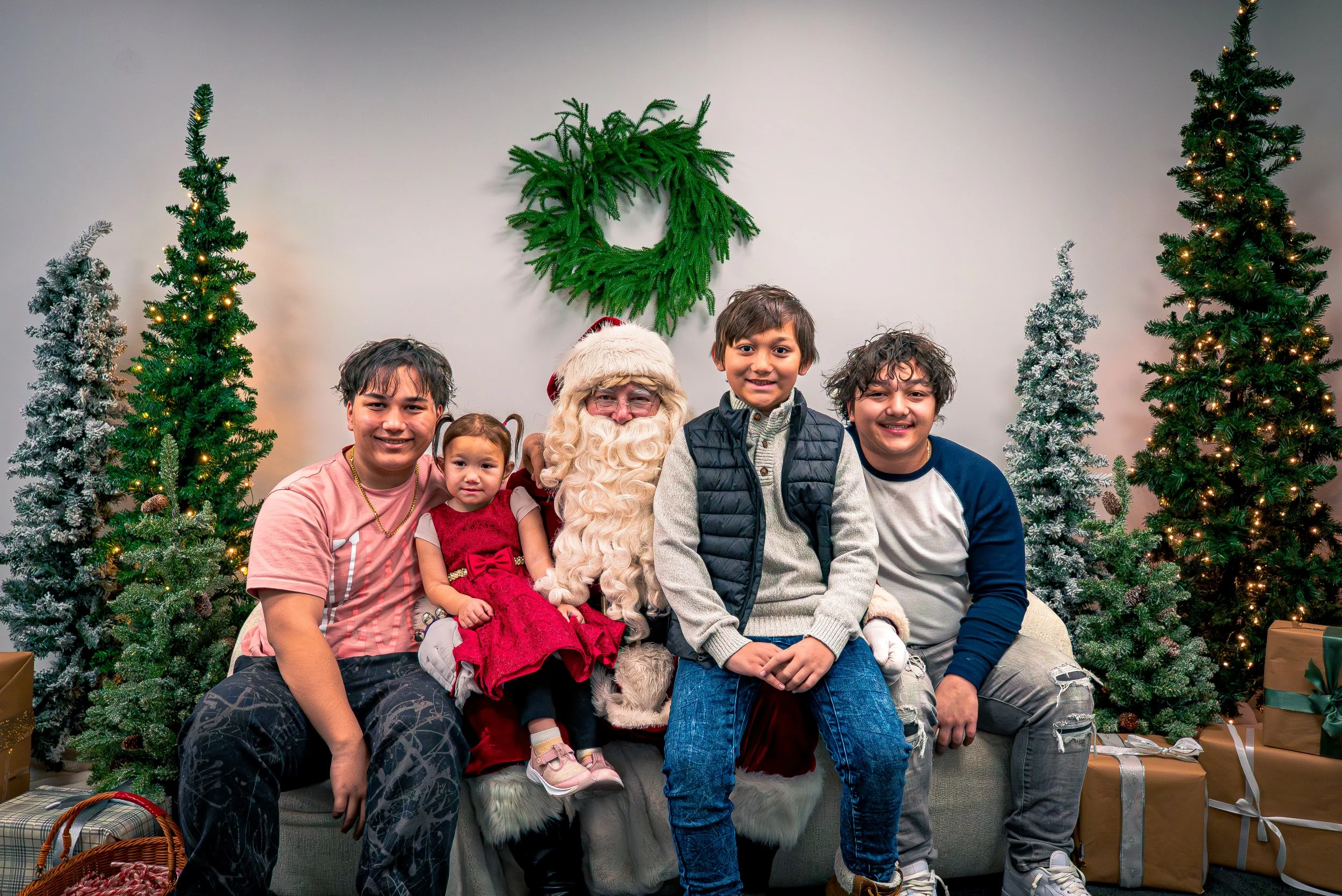 A group of five children and Santa Claus sitting together in front of a Christmas tree and a snowy decorated tree, posing for a holiday photo.