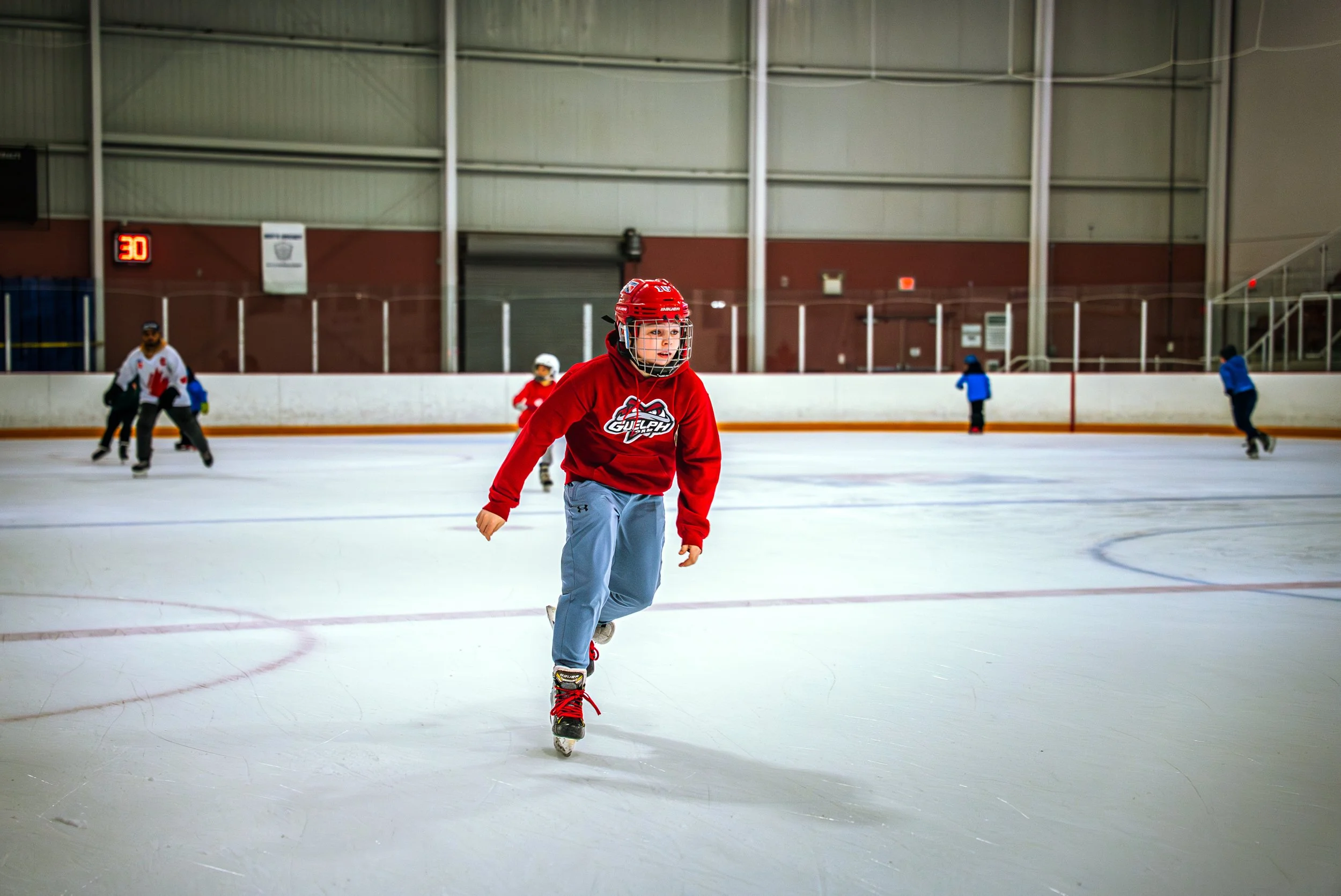 A young person skating on an indoor ice rink, wearing a red hockey helmet and a red sweatshirt with a team logo, surrounded by other skaters in the background.