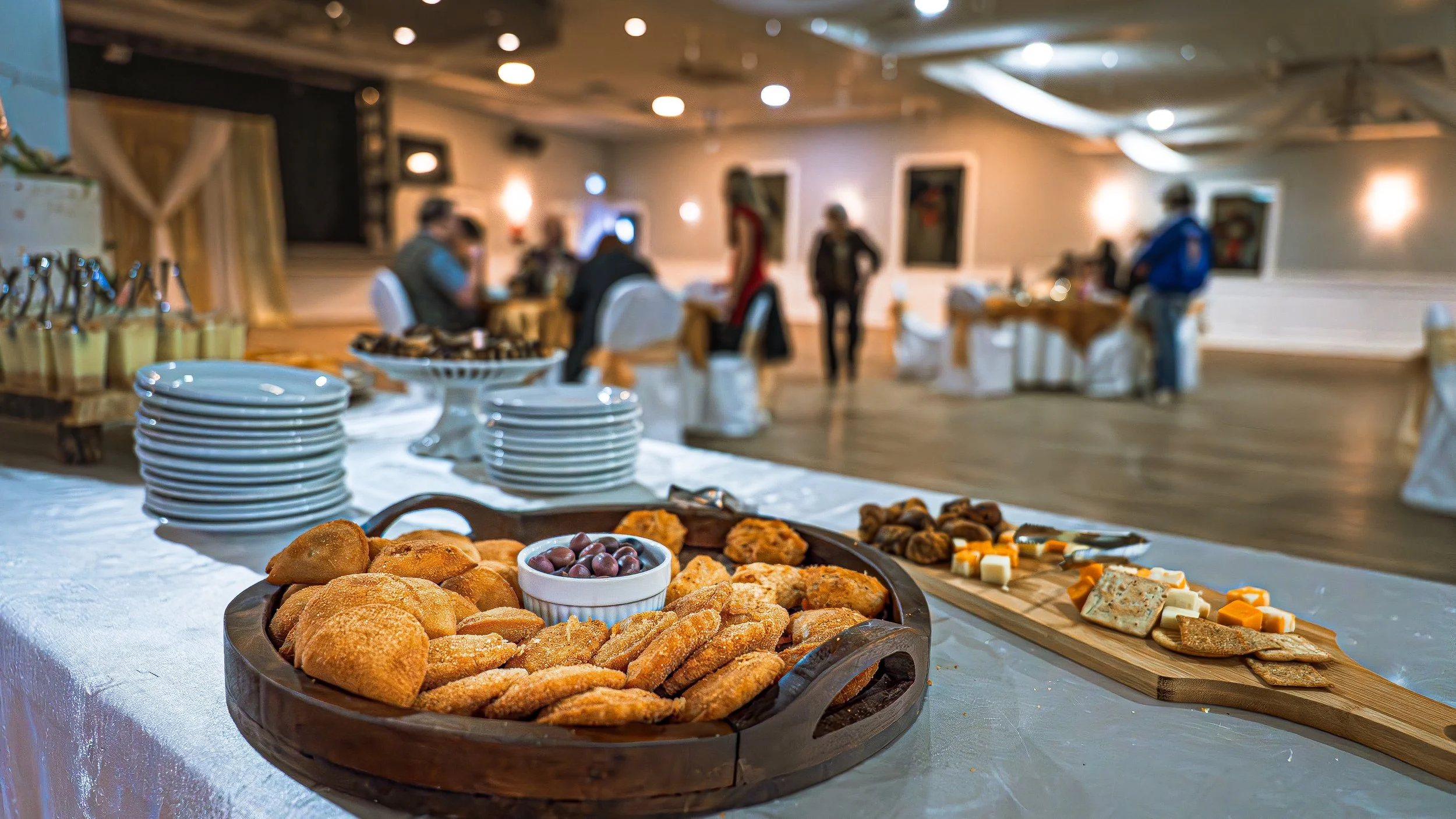Buffet table with assorted bread, cheese, olives, and snacks, with people in the background in a banquet hall.