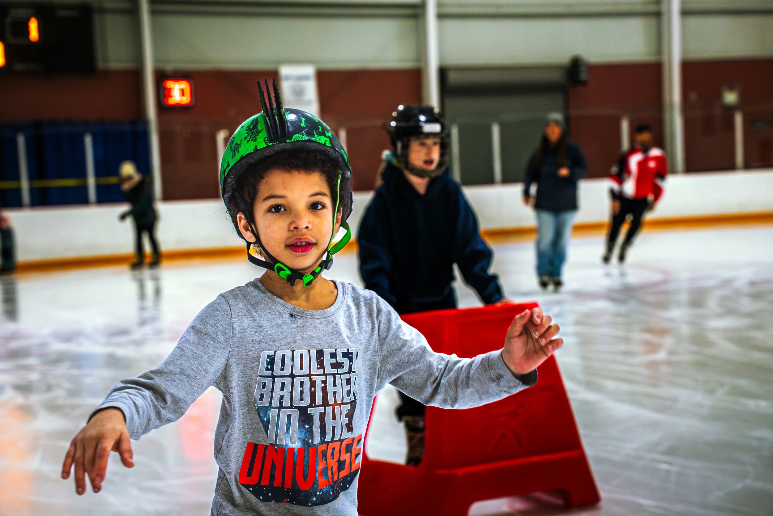 A young boy with curly hair wearing a helmet and a gray shirt with colorful text, standing on an ice rink. Other people are skating in the background, some with helmets, in a sports facility.
