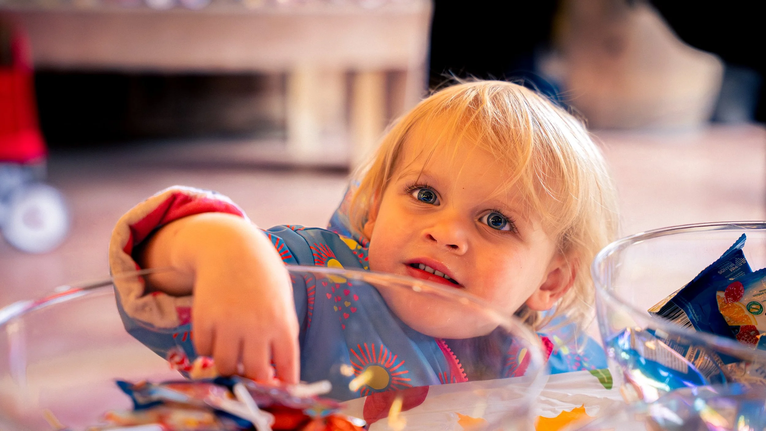 A young child with blond hair and blue eyes leaning over a clear glass bowl, reaching toward colorful snack packages, with a blurred background.