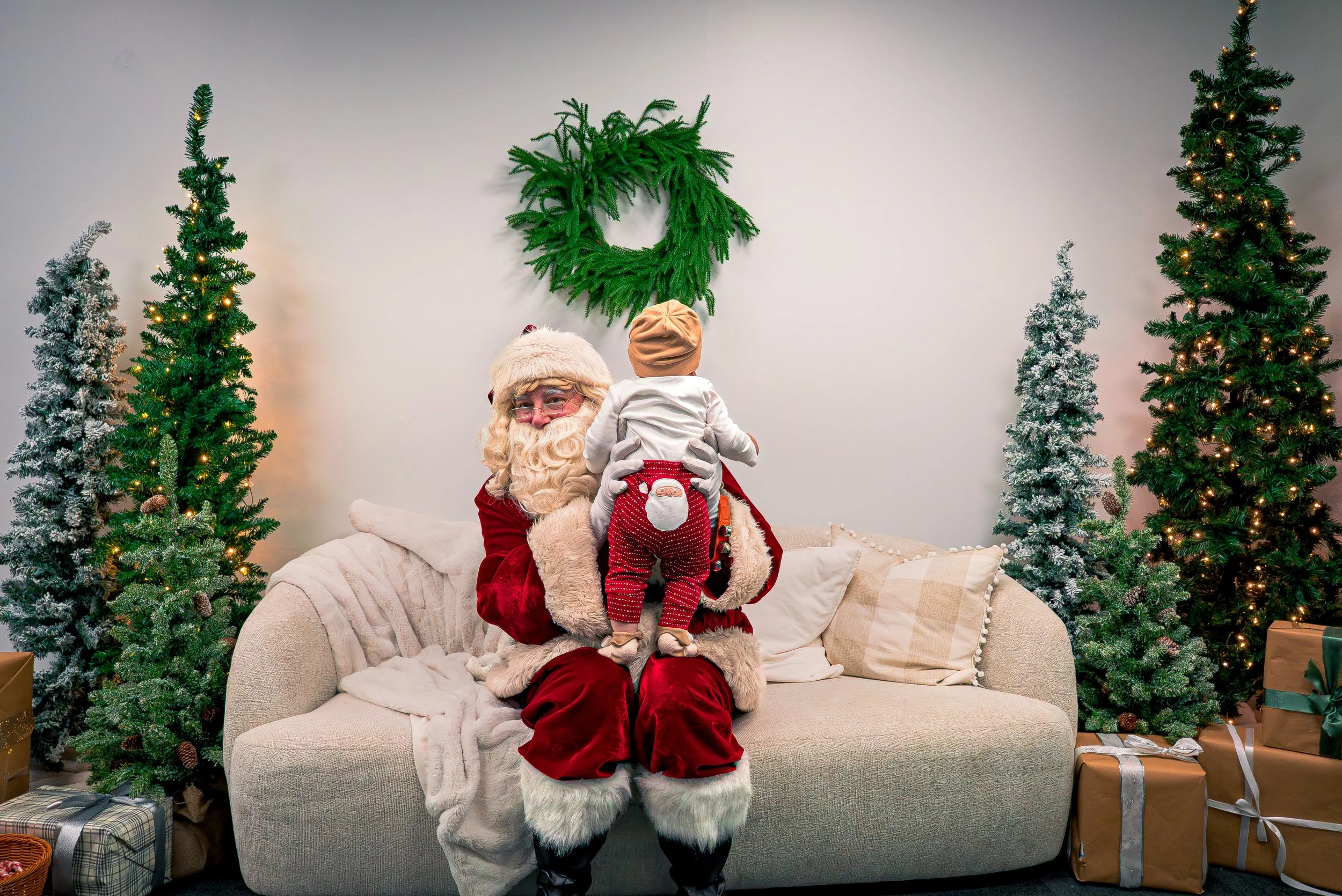 Santa Claus in a red suit with white fur trim holding a young child dressed in Christmas pajamas, surrounded by decorated Christmas trees and wrapped presents.