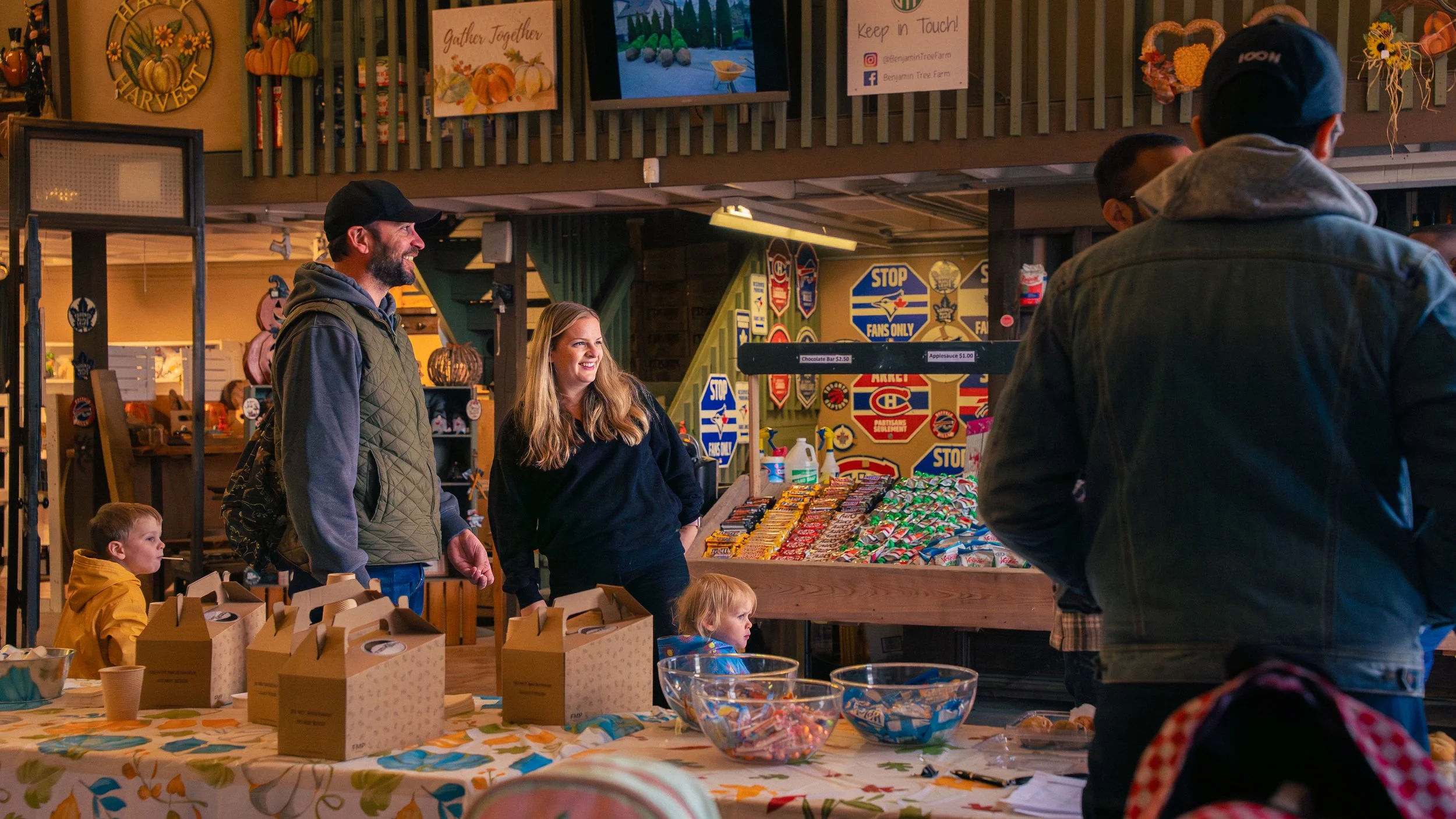 People shopping at an indoor market or store with souvenirs, candy, and signs for hockey and hockey teams. There are children and adults engaging with the vendor and browsing items.