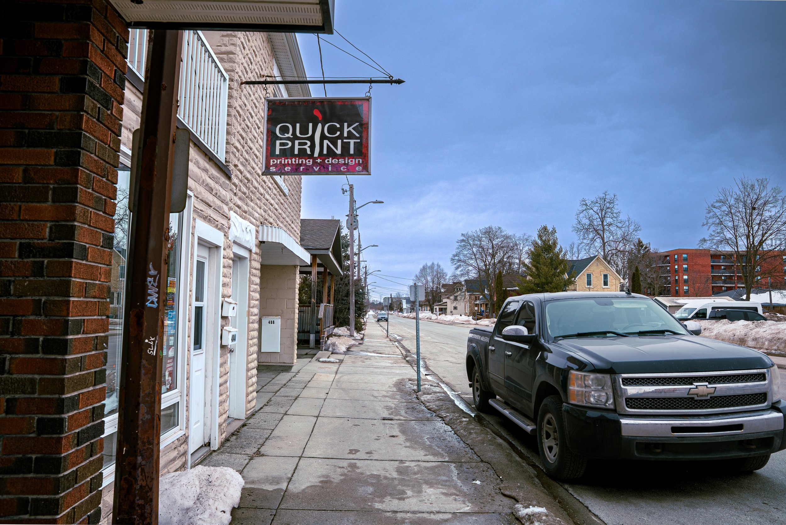 A street view with a business sign for 'Quick Print' hanging above the sidewalk. The sign indicates printing and design services. There are parked cars along the street, including a gray pickup truck, and several houses and trees in the background. S