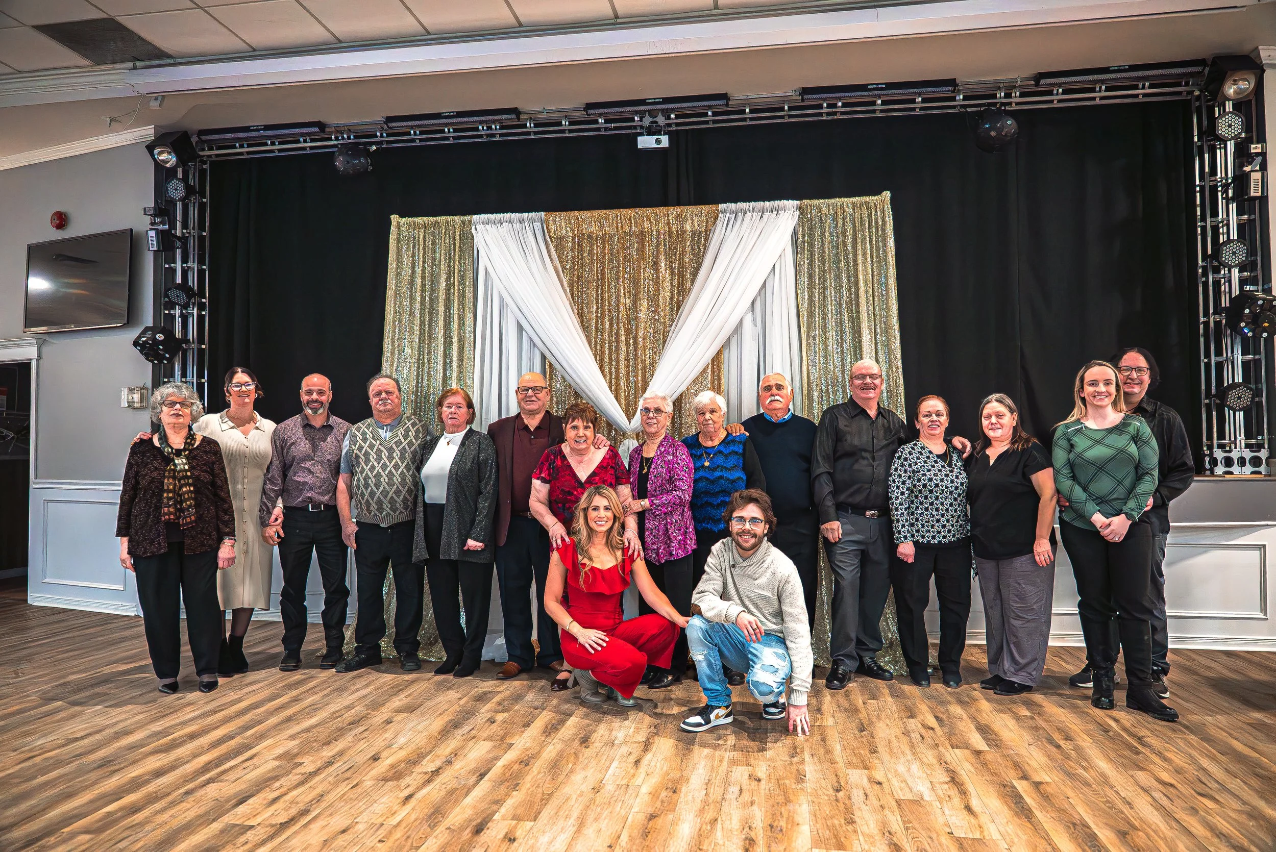 A large group of people of various ages posing for a photo in front of a decorated backdrop featuring gold and white curtains. The group appears to be at a celebration or event in an indoor venue with a black curtain and stage lighting.