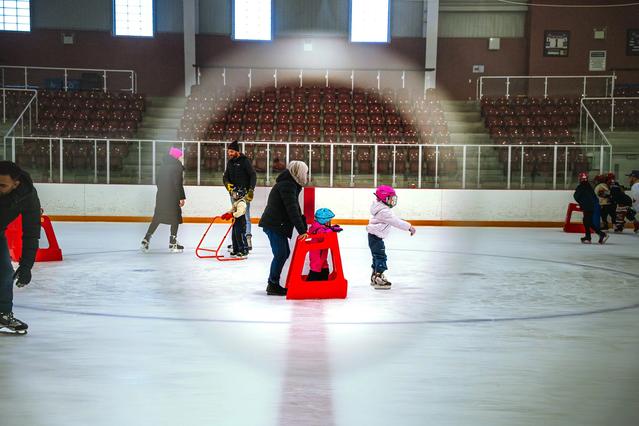 Children and adults ice skating at an indoor ice rink with empty red stadium seats in the background.