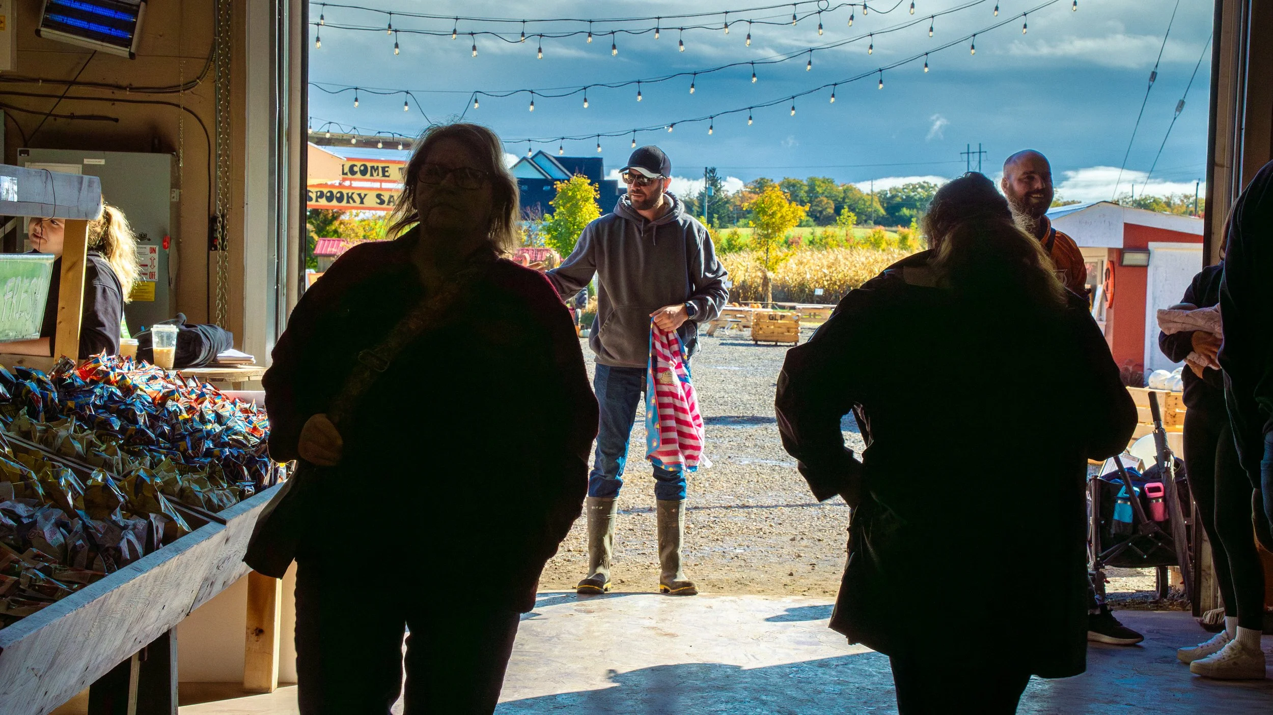 People shopping at a farm market, with some customers inside the barn and others outside under string lights, with a barn and fields in the background.
