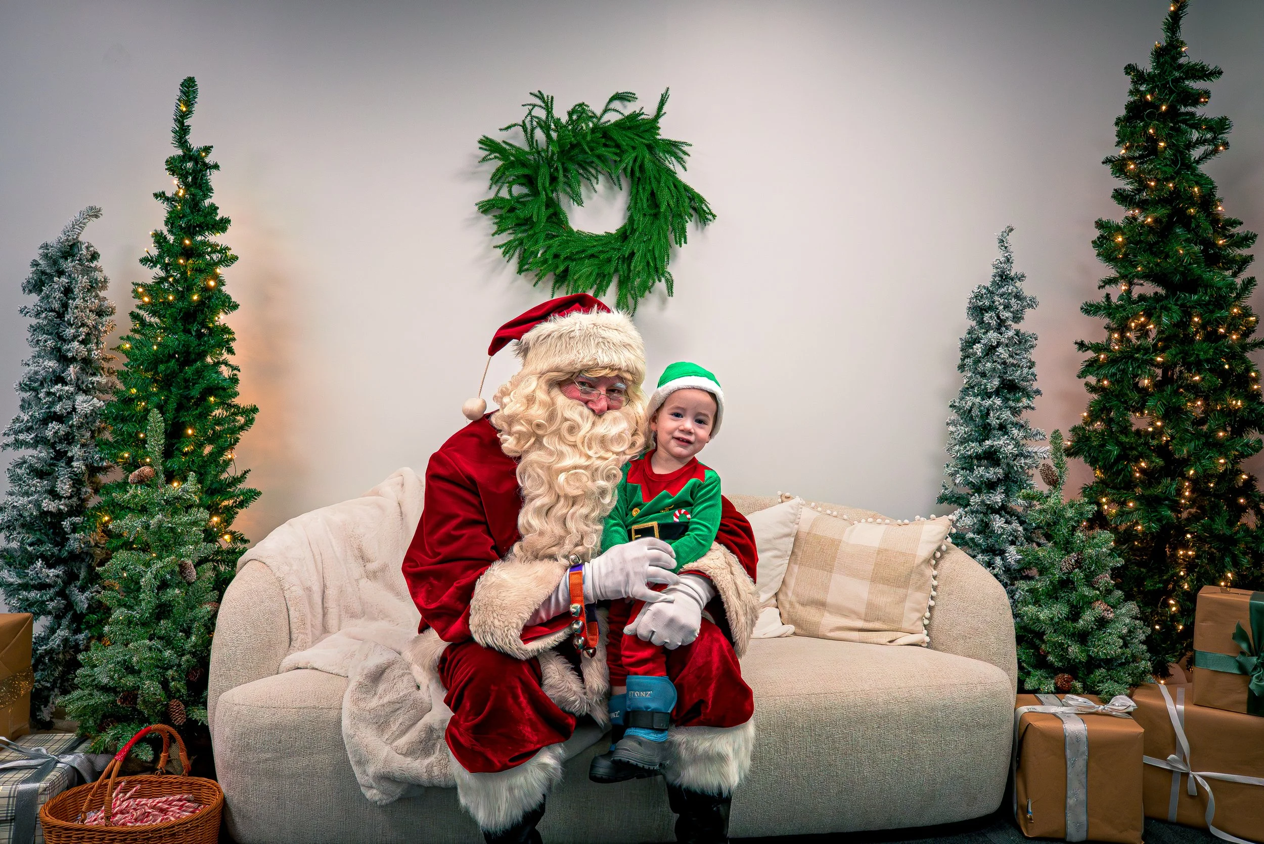A young child sitting on Santa's lap during a Christmas celebration. Santa is wearing a traditional red suit with white fur trim, glasses, and a Santa hat. The child is dressed in a festive Christmas outfit, smiling and looking at the camera. The bac