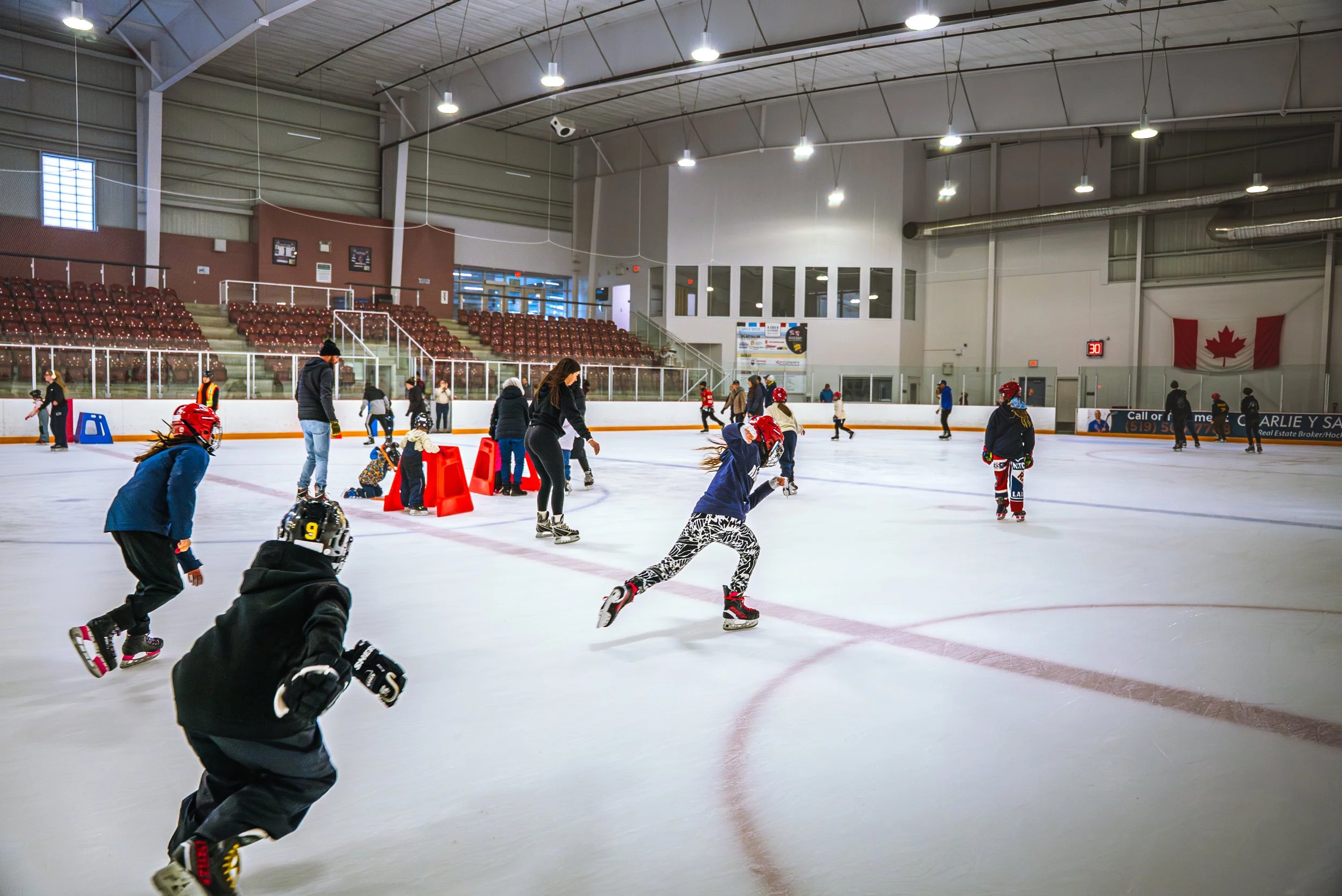 People ice skating at an indoor rink with overhead lights, seating area, and Canadian flag.