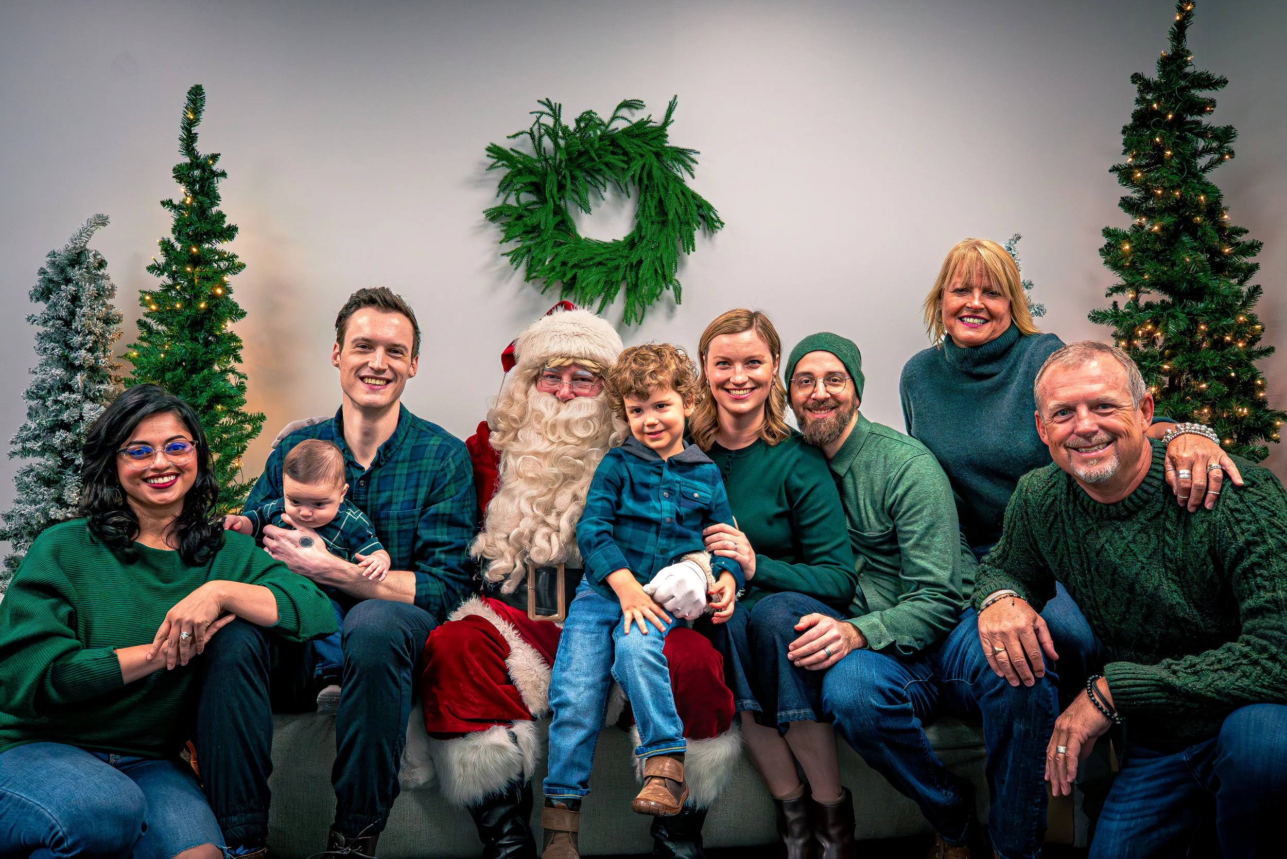 A group of people including children, adults, and Santa Claus, gathered together smiling in front of a plain wall decorated with Christmas trees and a lush green wreath.