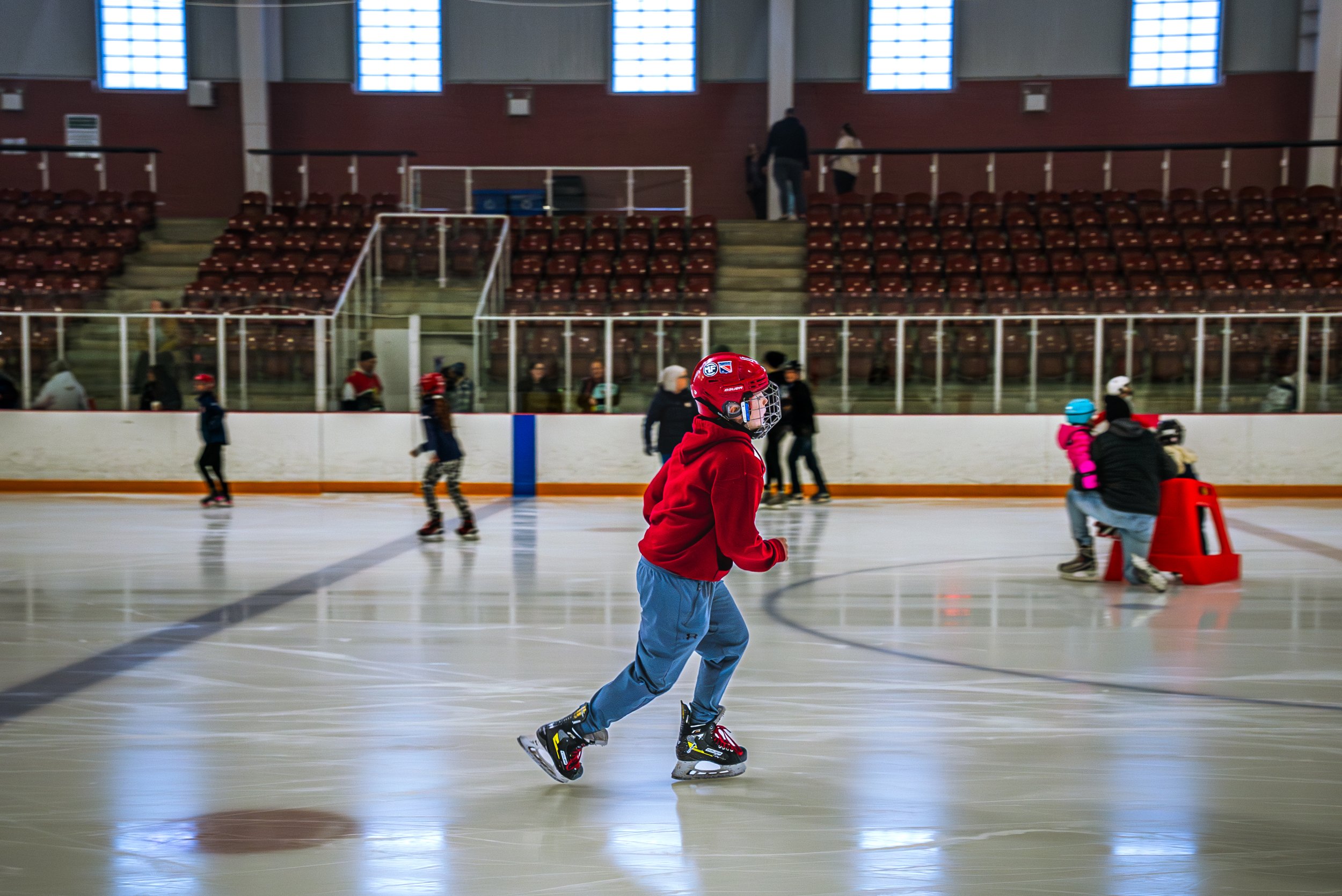 A young person in a red hoodie and blue pants roller skating inside an ice rink, with several other skaters in the background, and empty spectator seats above.