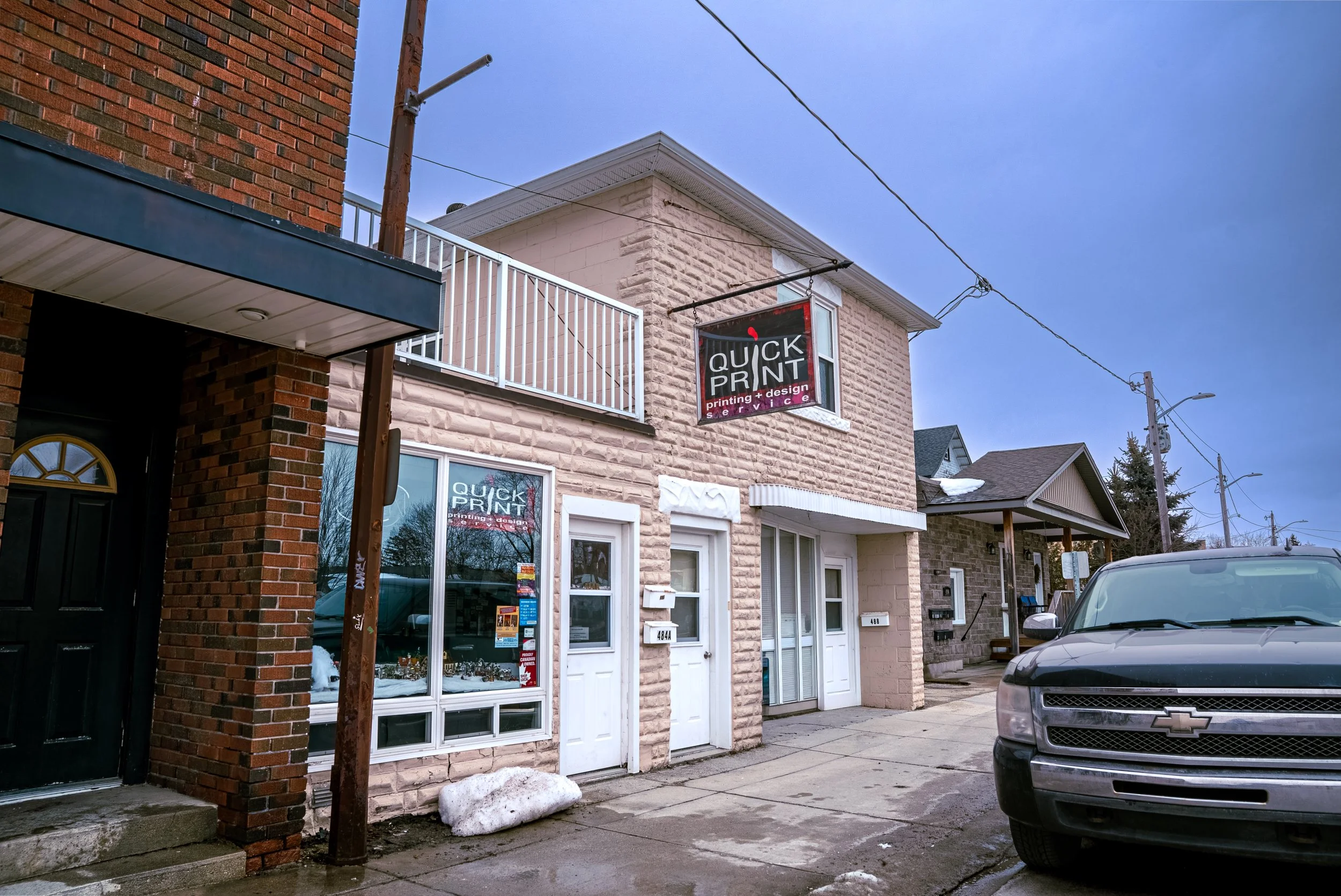 Street view of a building with a sign for 'Quick Print' printing and design service, with residential houses and parked cars in the background, and patches of snow on the sidewalk.