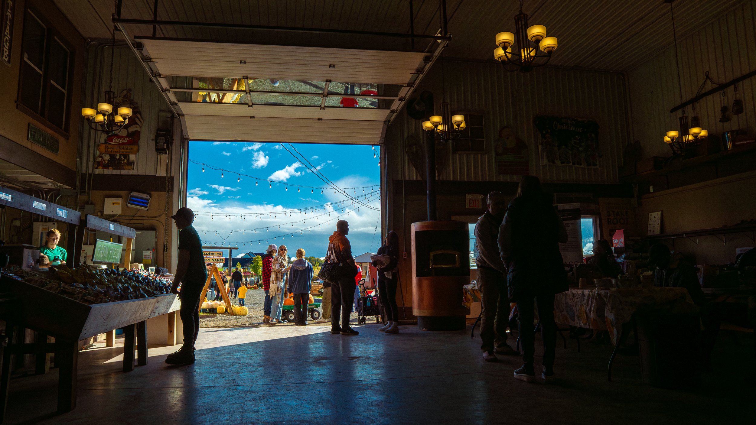 View from inside a barn or covered area looking out onto a busy outdoor market with people shopping and walking, with bright blue sky and clouds above.