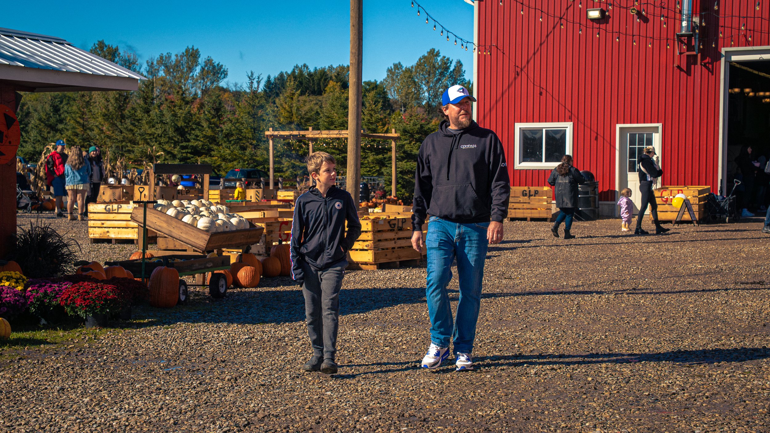 People walking outside a farm stand with pumpkins and fall flowers on display, red barn in the background, with trees and a blue sky.