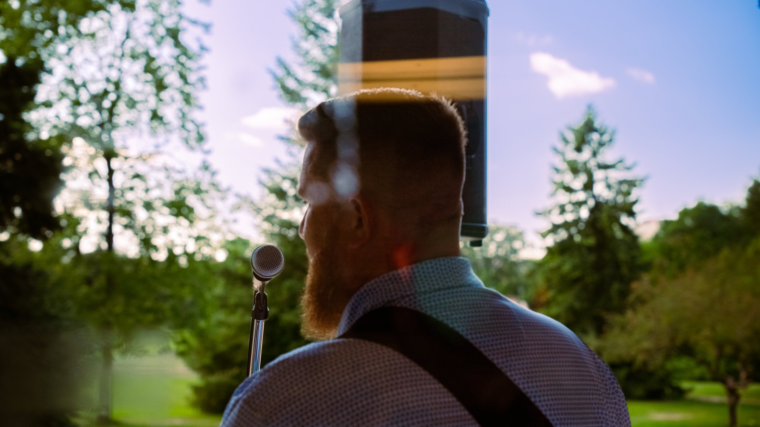 A man with short hair and a beard wearing a collared shirt, standing outside in front of trees and blue sky, seen through a reflective glass window.