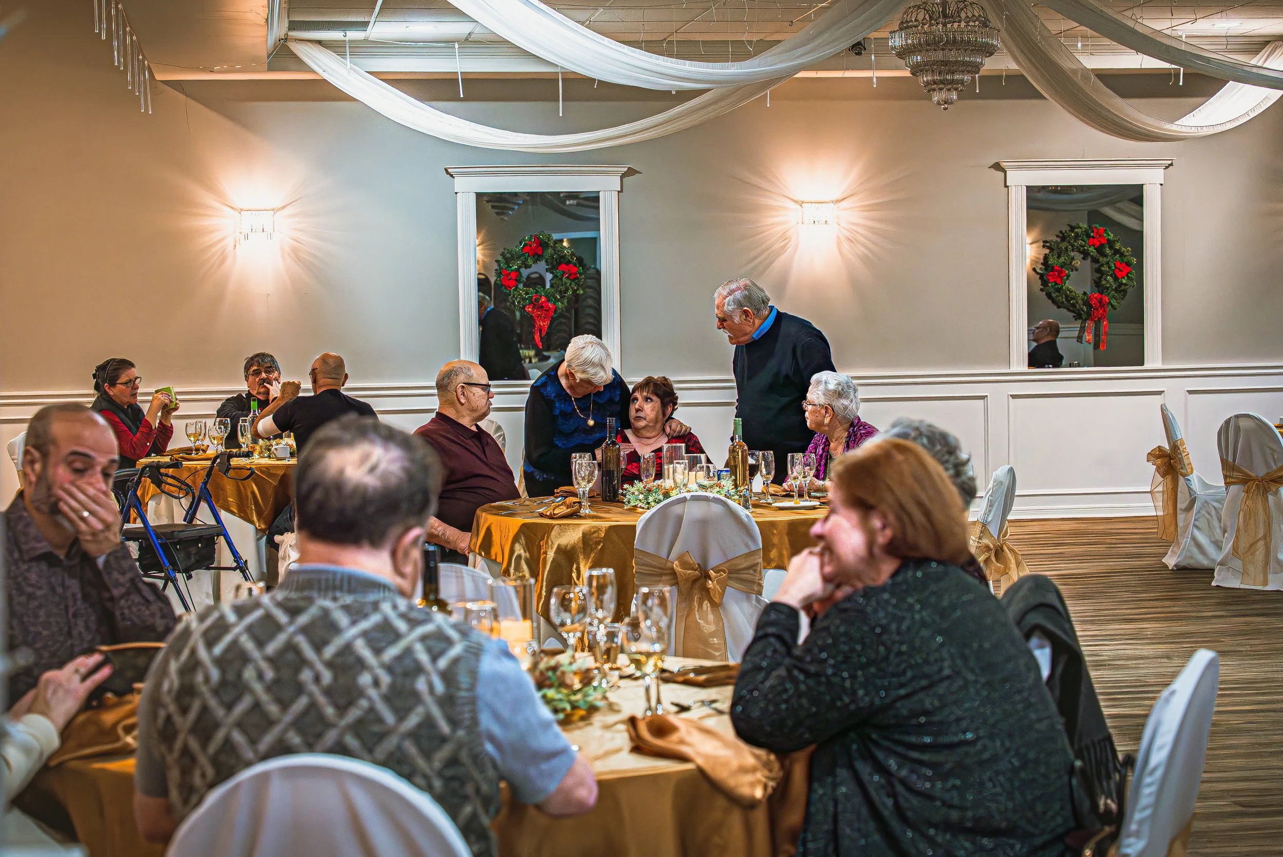 People gathered at a Christmas celebration in a decorated dining hall, with wreaths and draped fabric on the ceiling.